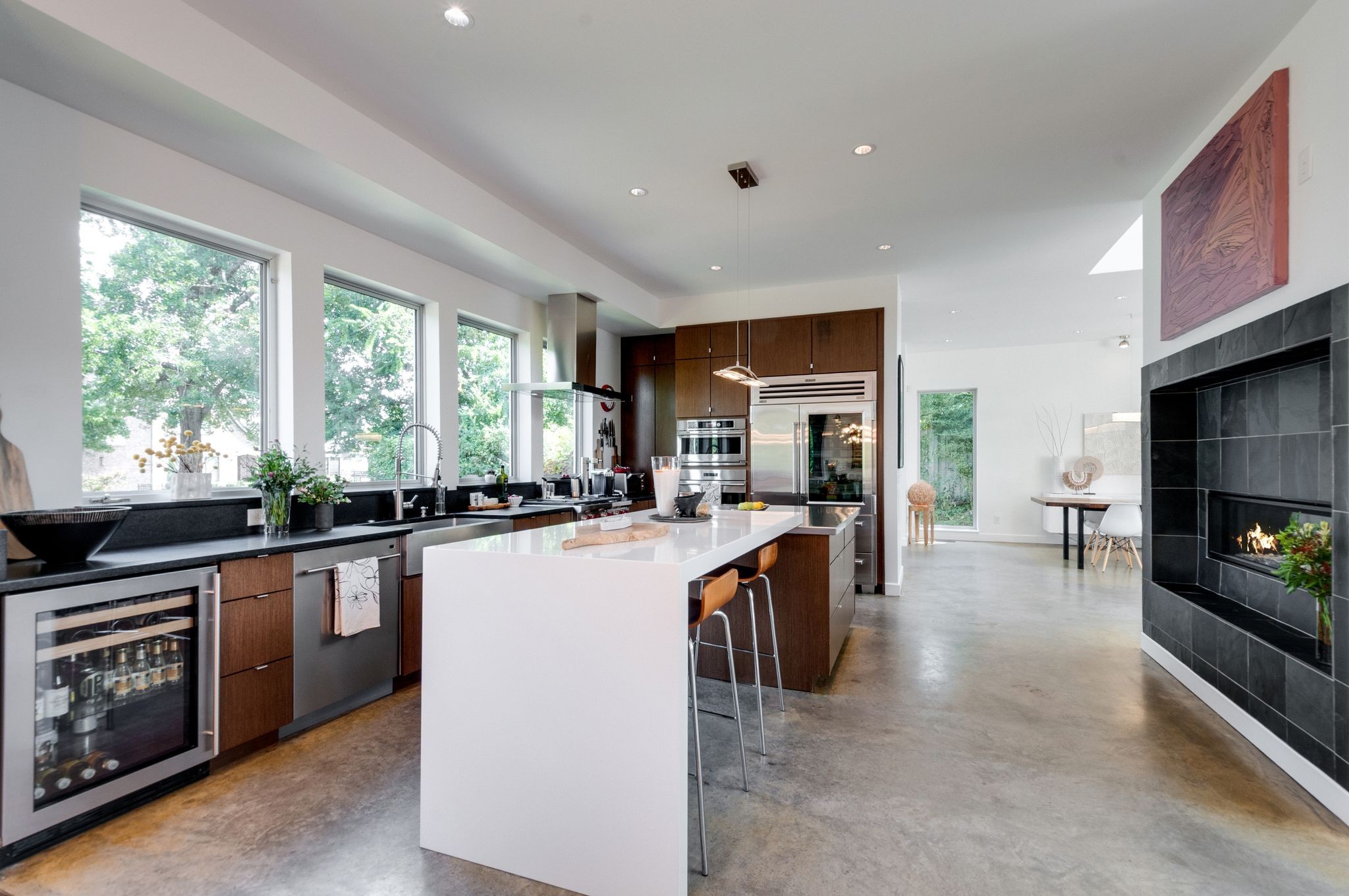 Modern kitchen with a large white island, tall windows, dark cabinetry, and an adjoining fireplace.