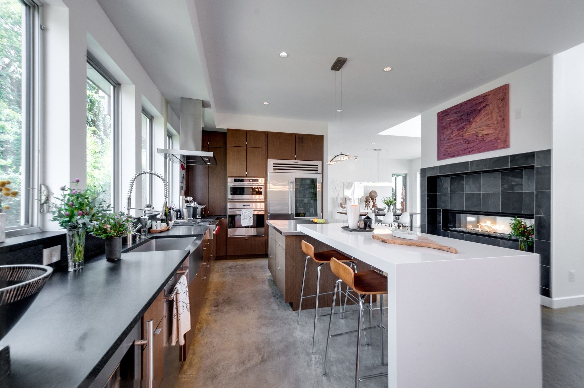 Modern kitchen with a large white island, tall windows, and a black-tiled fireplace.