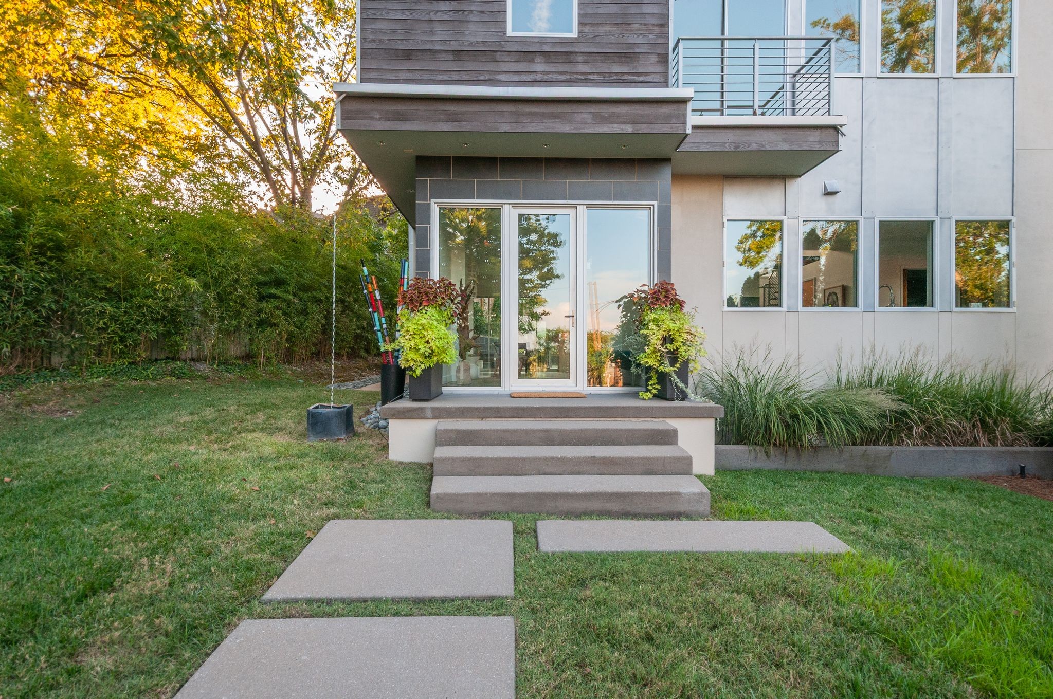 Modern home rear entrance with concrete steps, glass doors, an upper balcony, and a landscaped lawn.