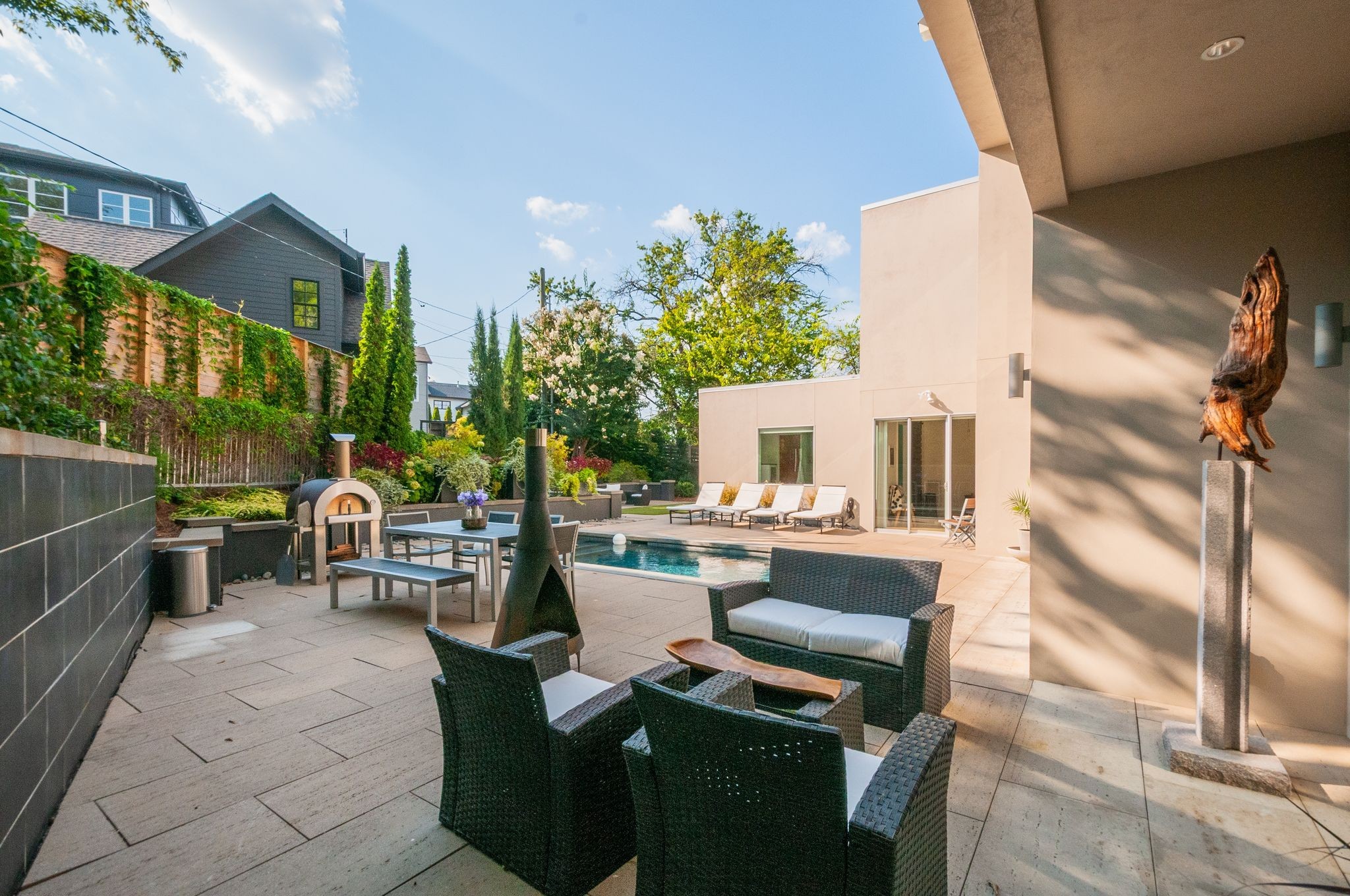 Modern home patio with pool, wicker seating, and a pizza oven beside the landscaped garden.