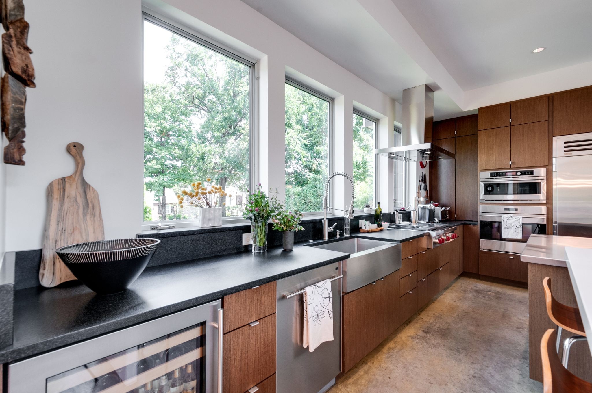 Modern kitchen with wood cabinetry, dark counters, stainless appliances, and tall windows overlooking trees.