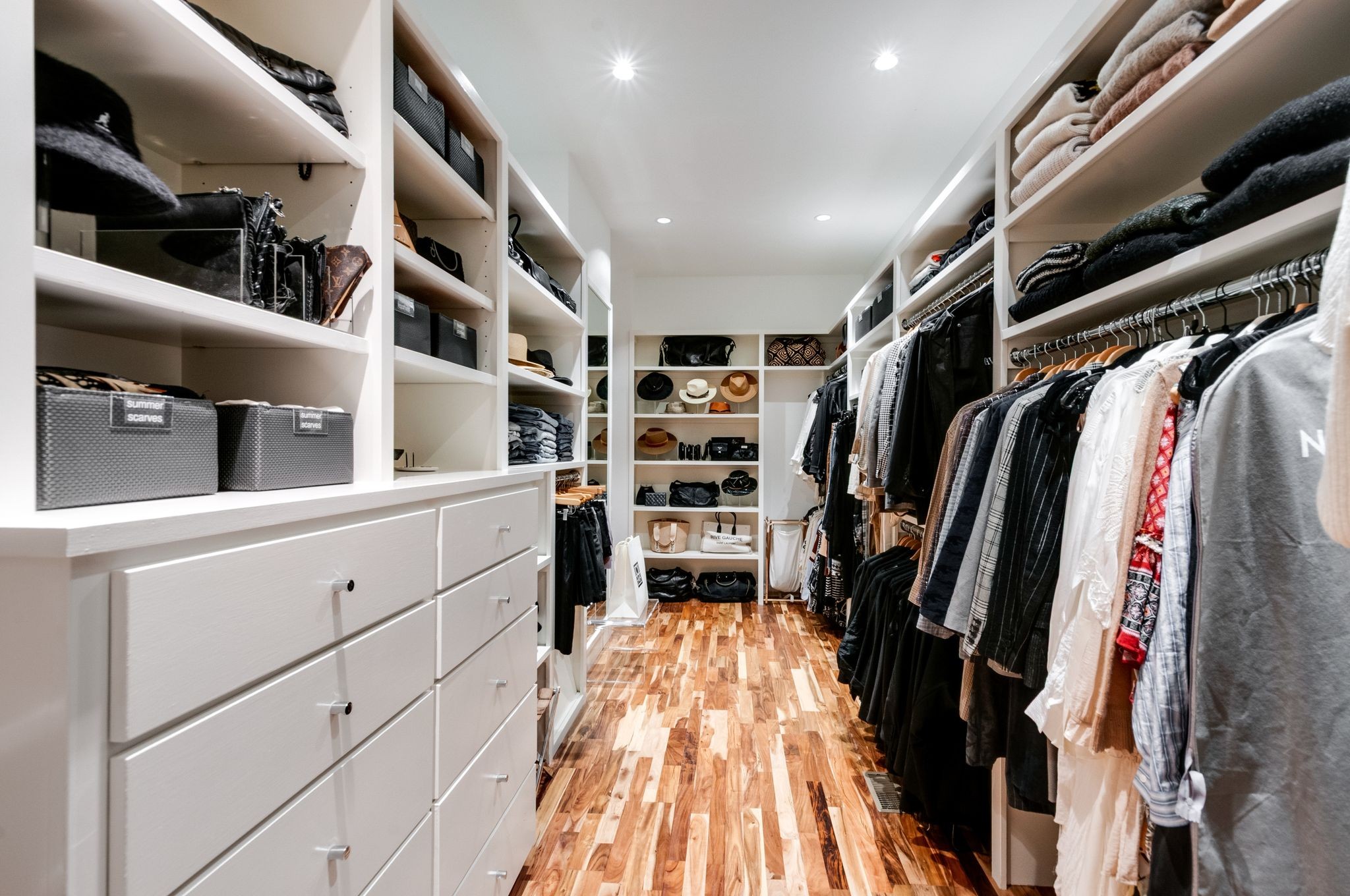 Walk-in closet with white built-ins, organized clothing, accessory shelves, and warm wood flooring.