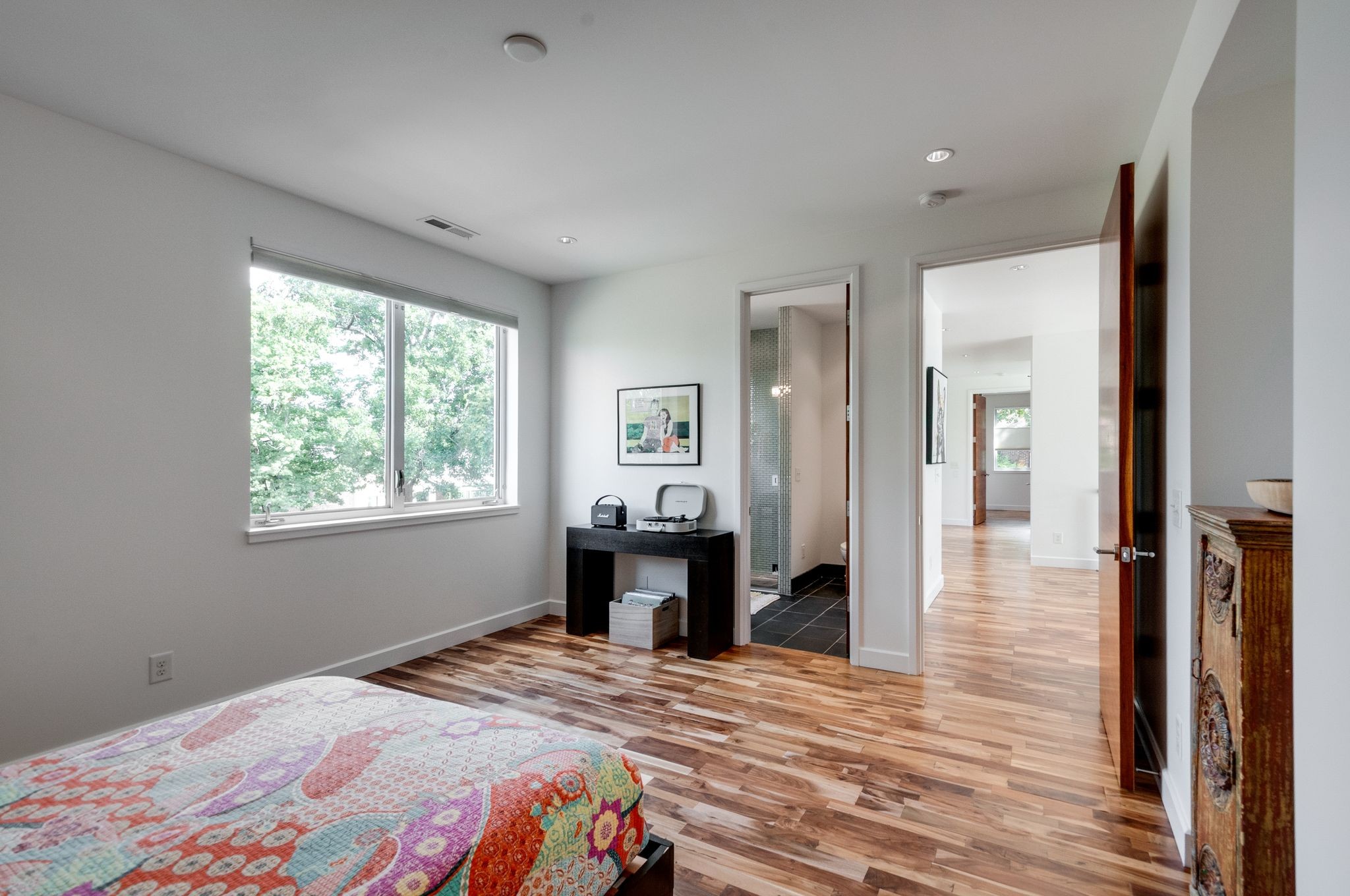 Bedroom with colorful patterned bedspread, hardwood floors, and a large window overlooking leafy trees.