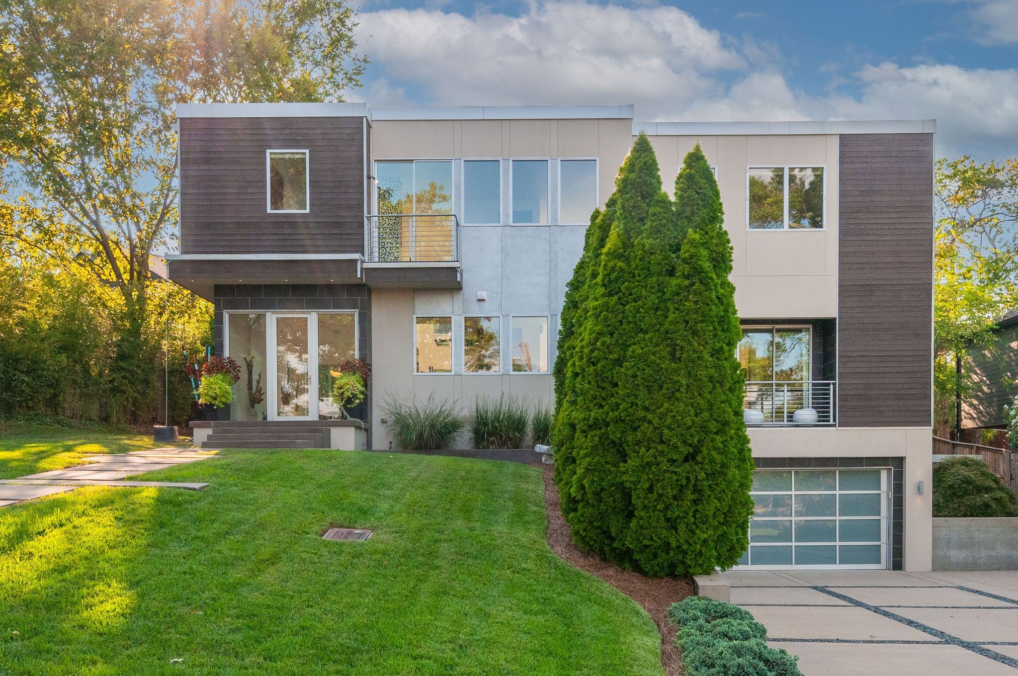 Modern two-story home with a balcony, attached garage, and tall evergreen in front.