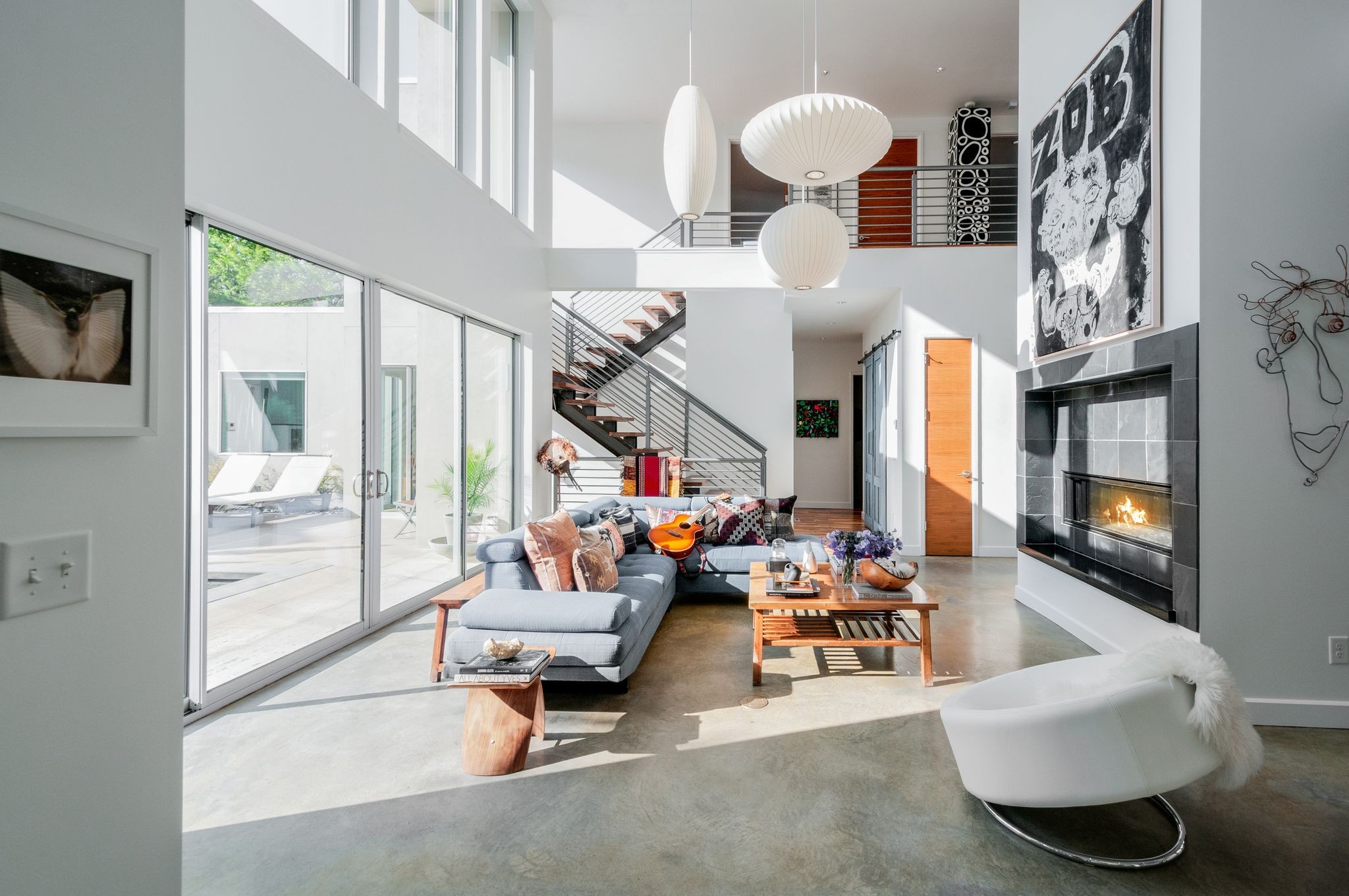 Double-height living room with a gray sectional, tall windows, open staircase, and lit fireplace.