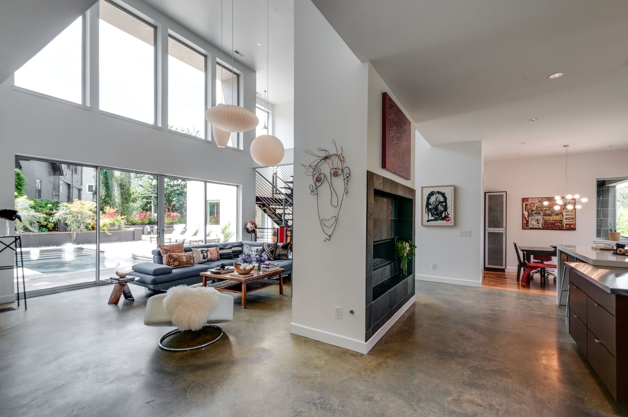 Open-plan living room with double-height windows, gray sectional, tiled fireplace, and views of a patio pool.