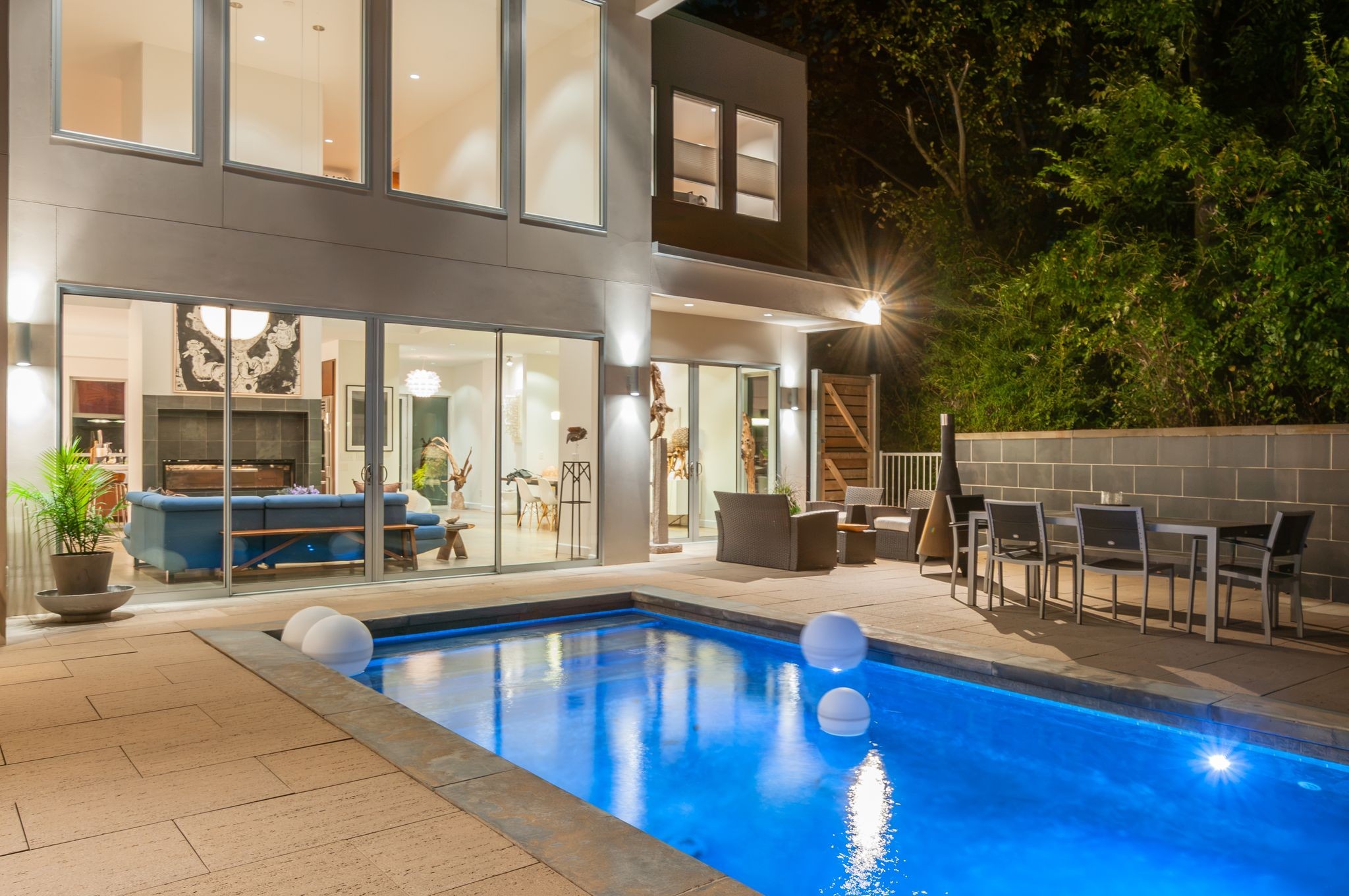 Modern home with a glowing swimming pool, glass walls, and furnished patio at night.