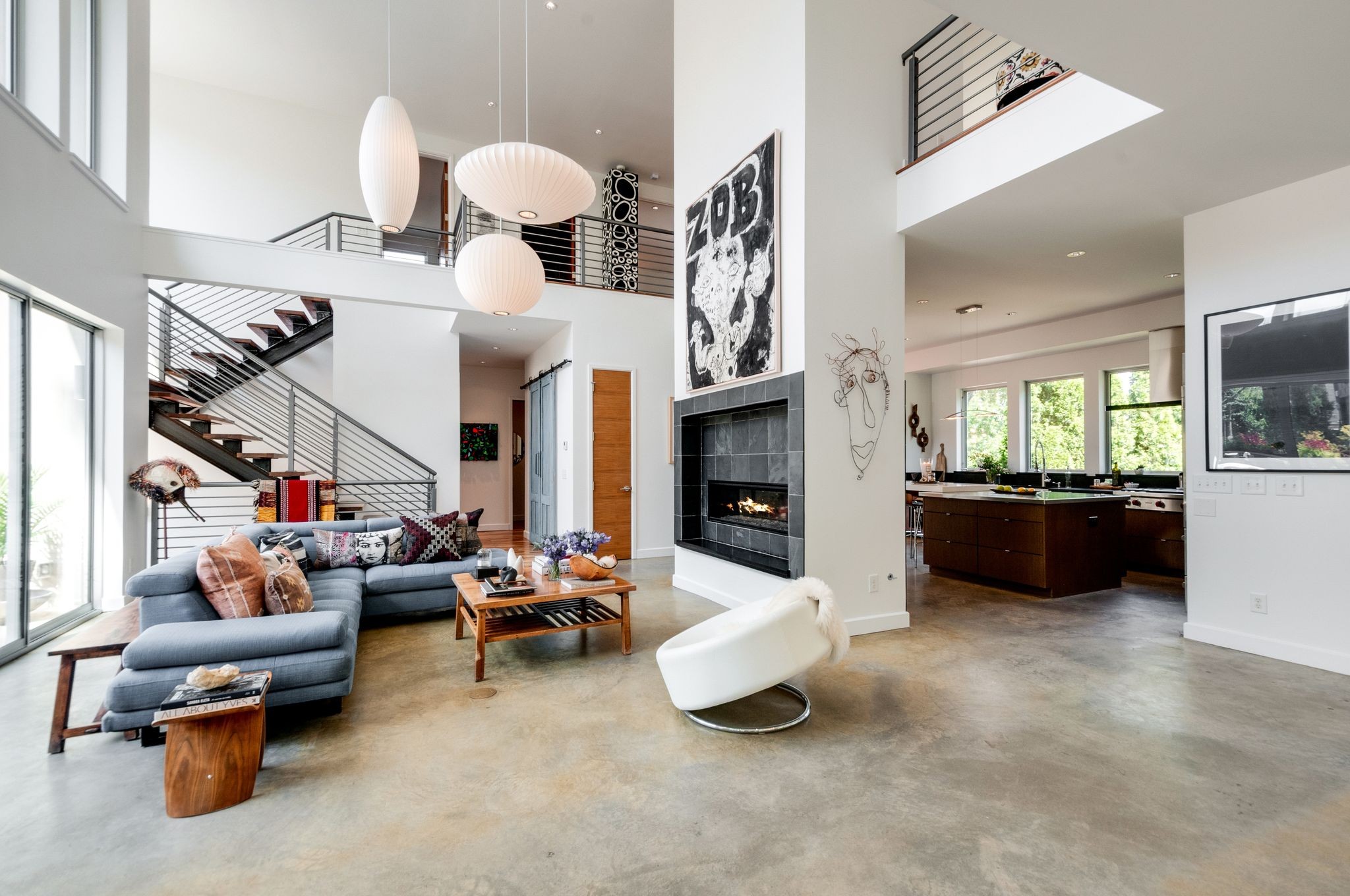 Open-plan living room with a double-height ceiling, metal-railed staircase, fireplace, and adjoining kitchen.