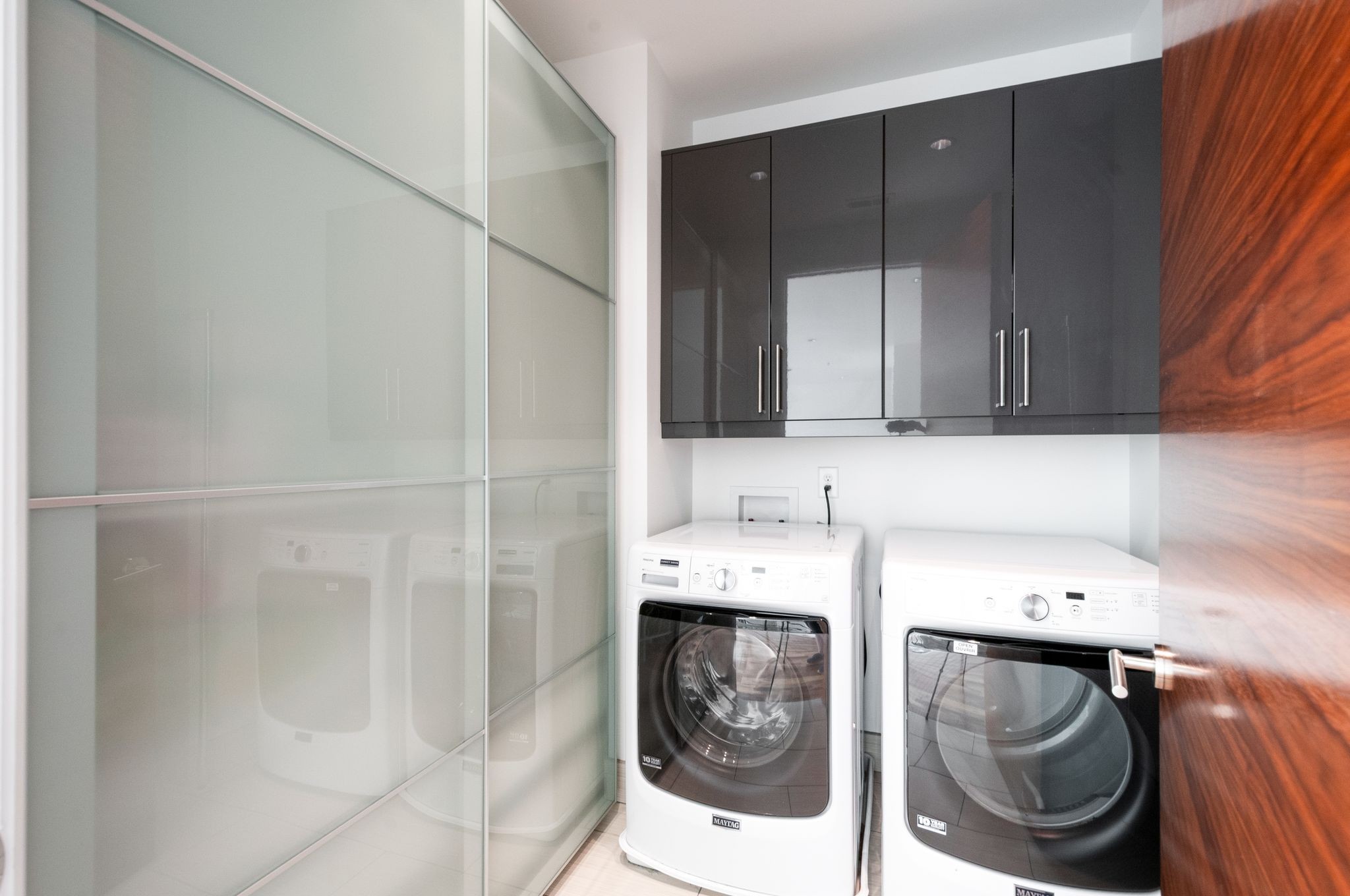 Laundry room with front-loading washer and dryer, dark cabinets, and a frosted glass sliding door.