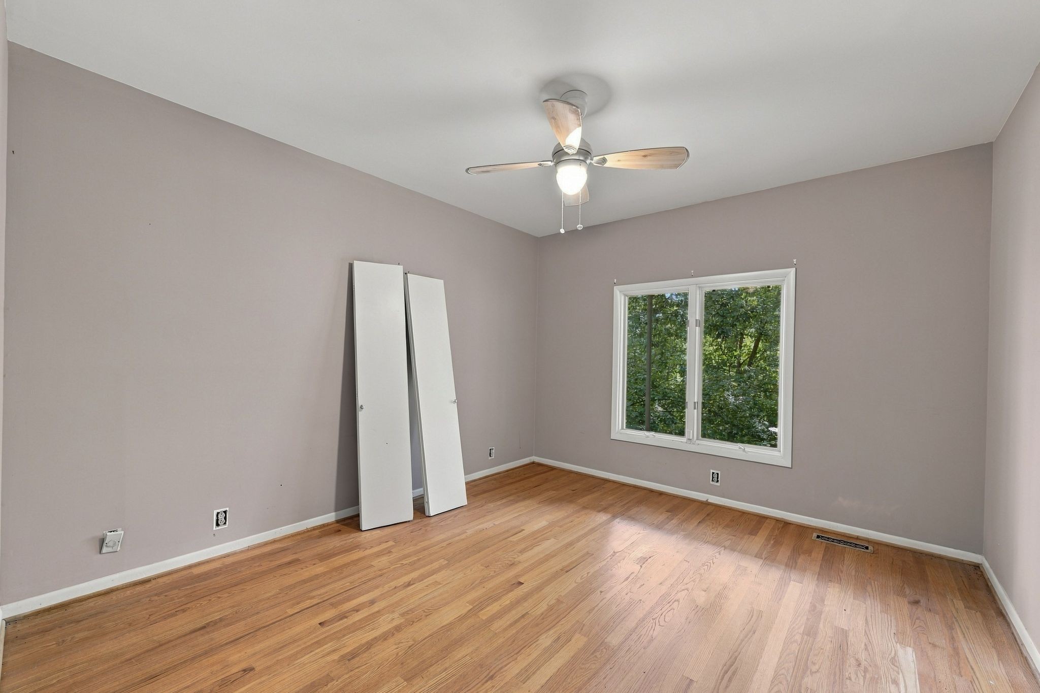 Empty bedroom with hardwood floors, a ceiling fan, and a window overlooking trees.
