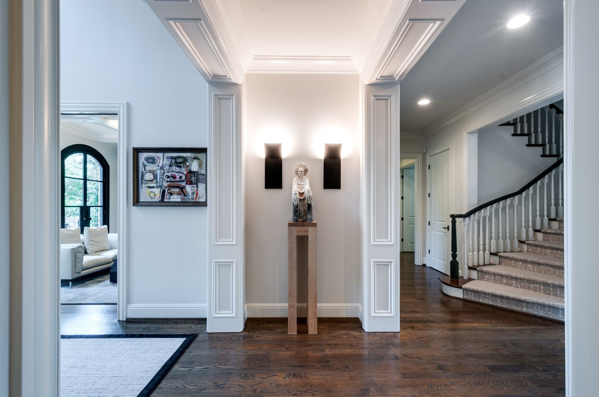 Home foyer with a statue beneath wall sconces, dark wood floors, and a carpeted staircase.