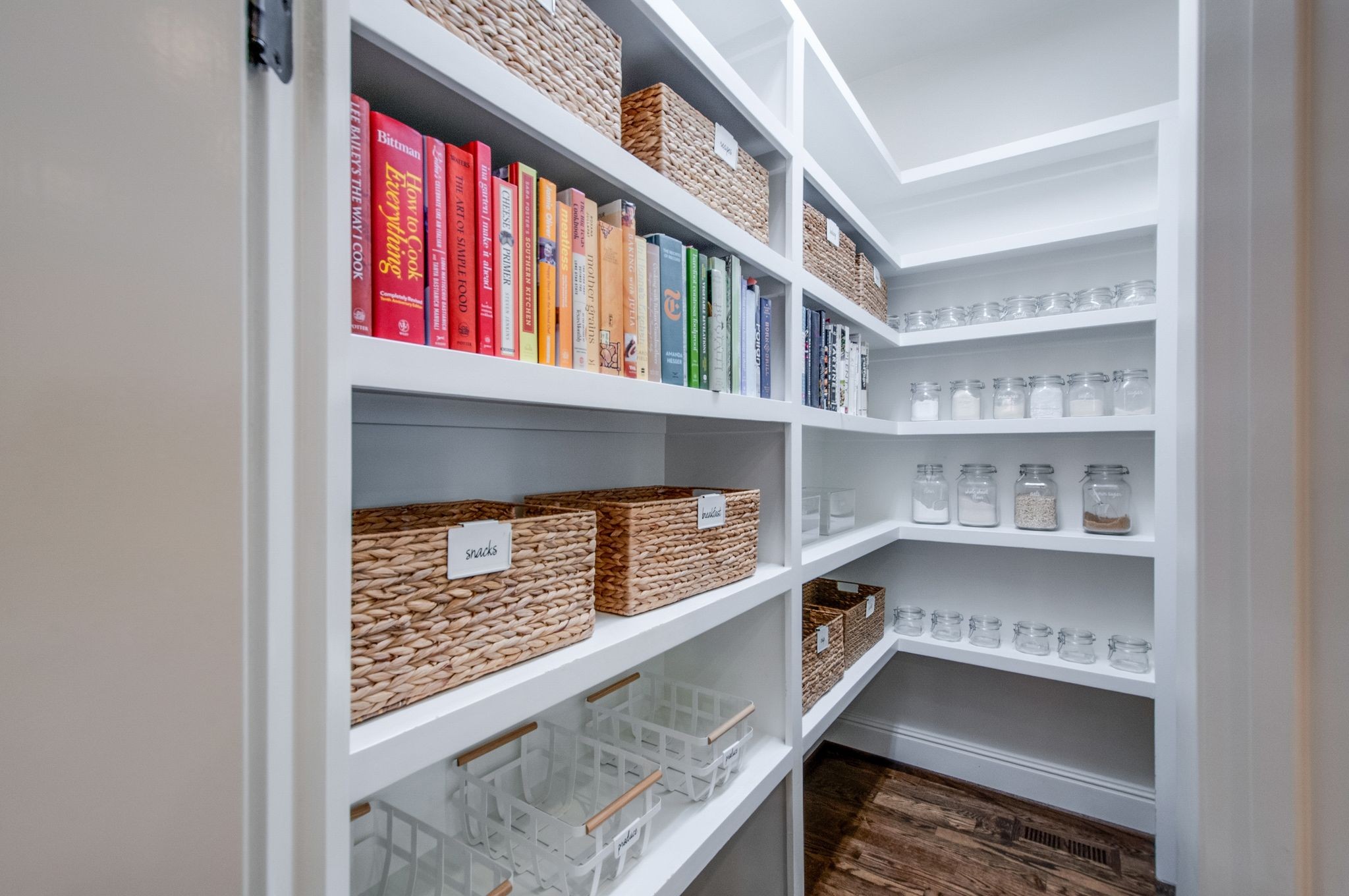 Walk-in pantry with white shelves, woven baskets, cookbooks, glass jars, and dark wood flooring.