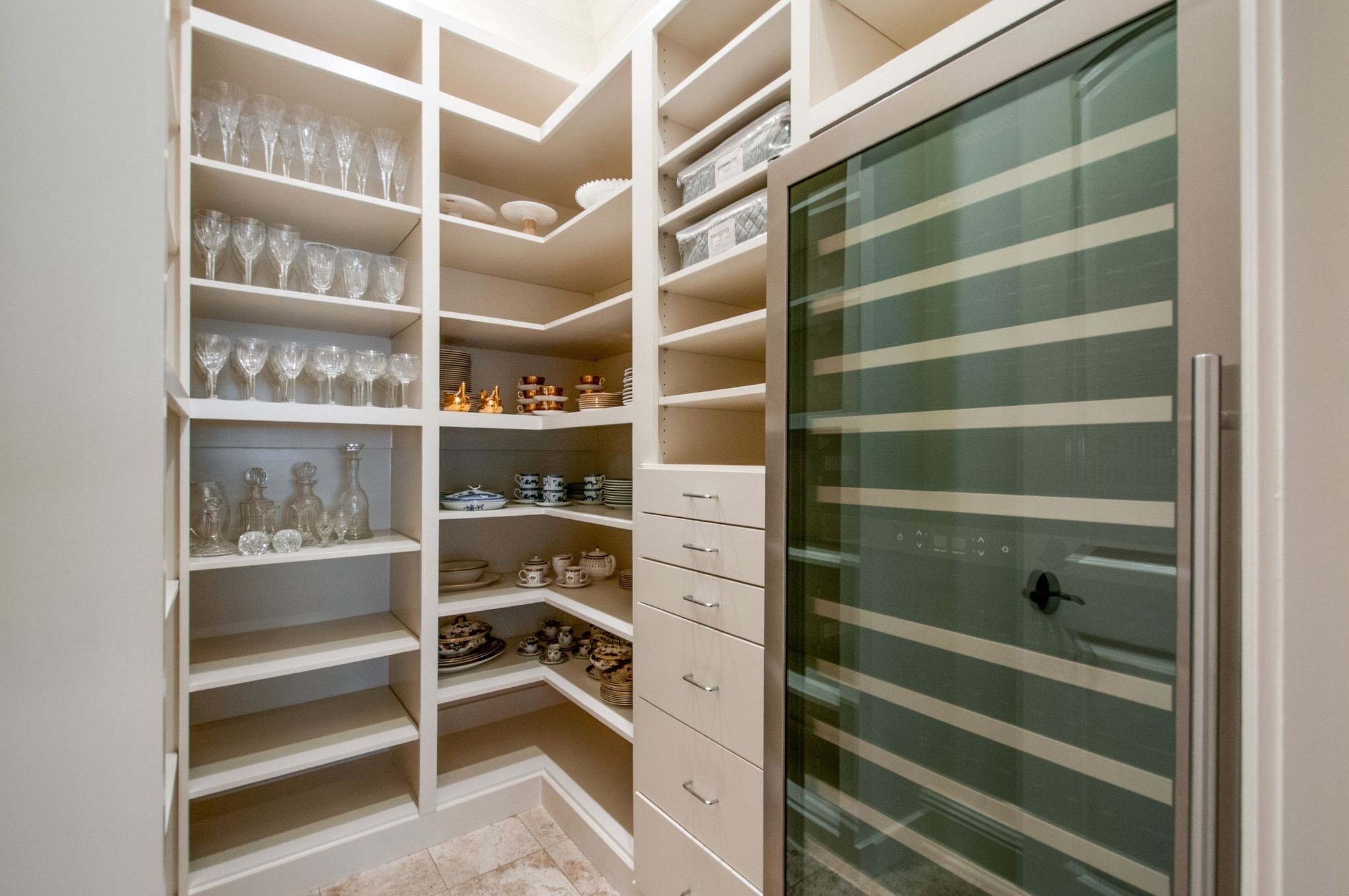 Walk-in pantry with built-in shelves of glassware and dishes beside a glass-front wine refrigerator.