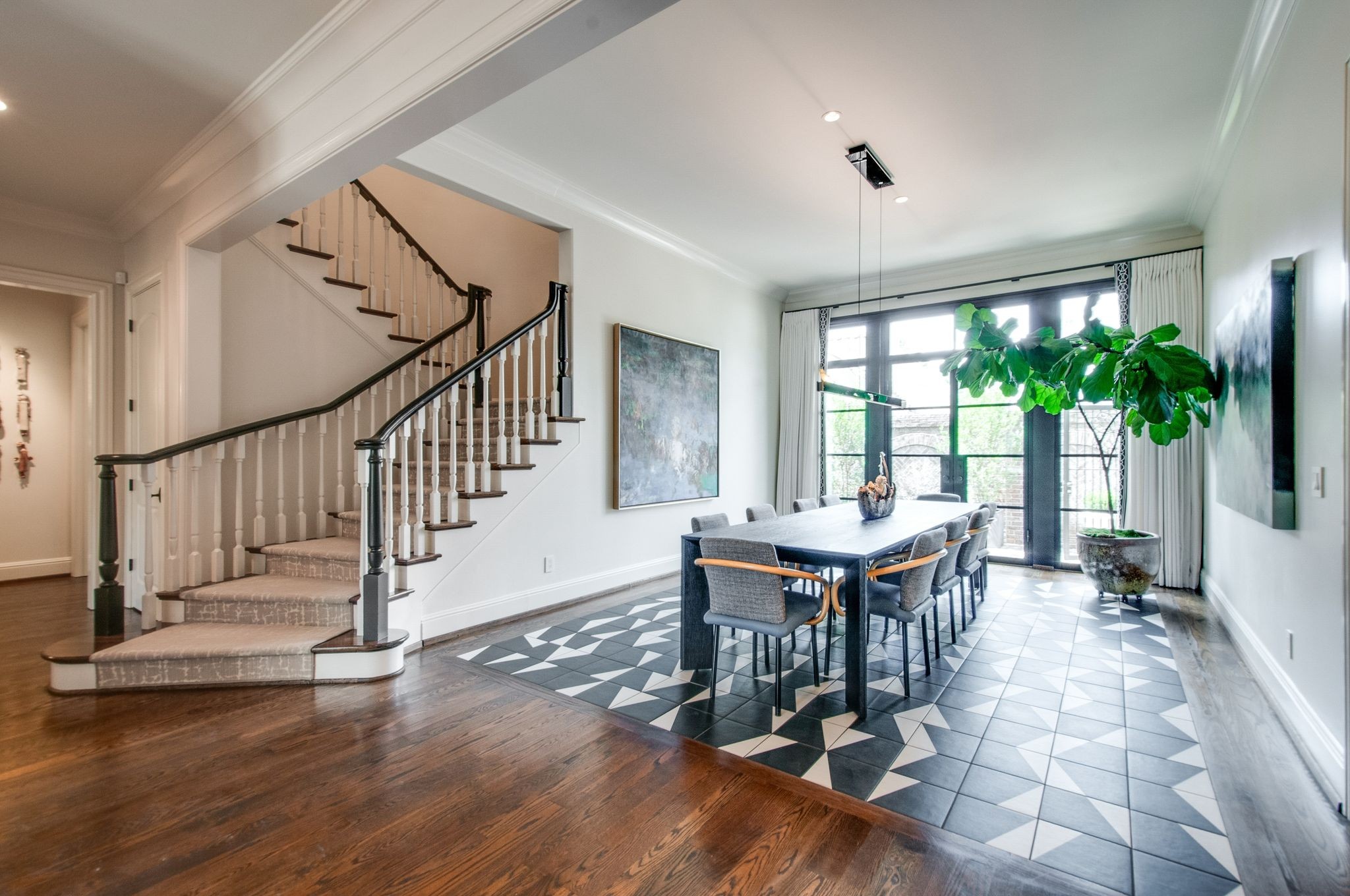 Dining room with geometric tile flooring, a long table, and a sweeping staircase beside large windows.