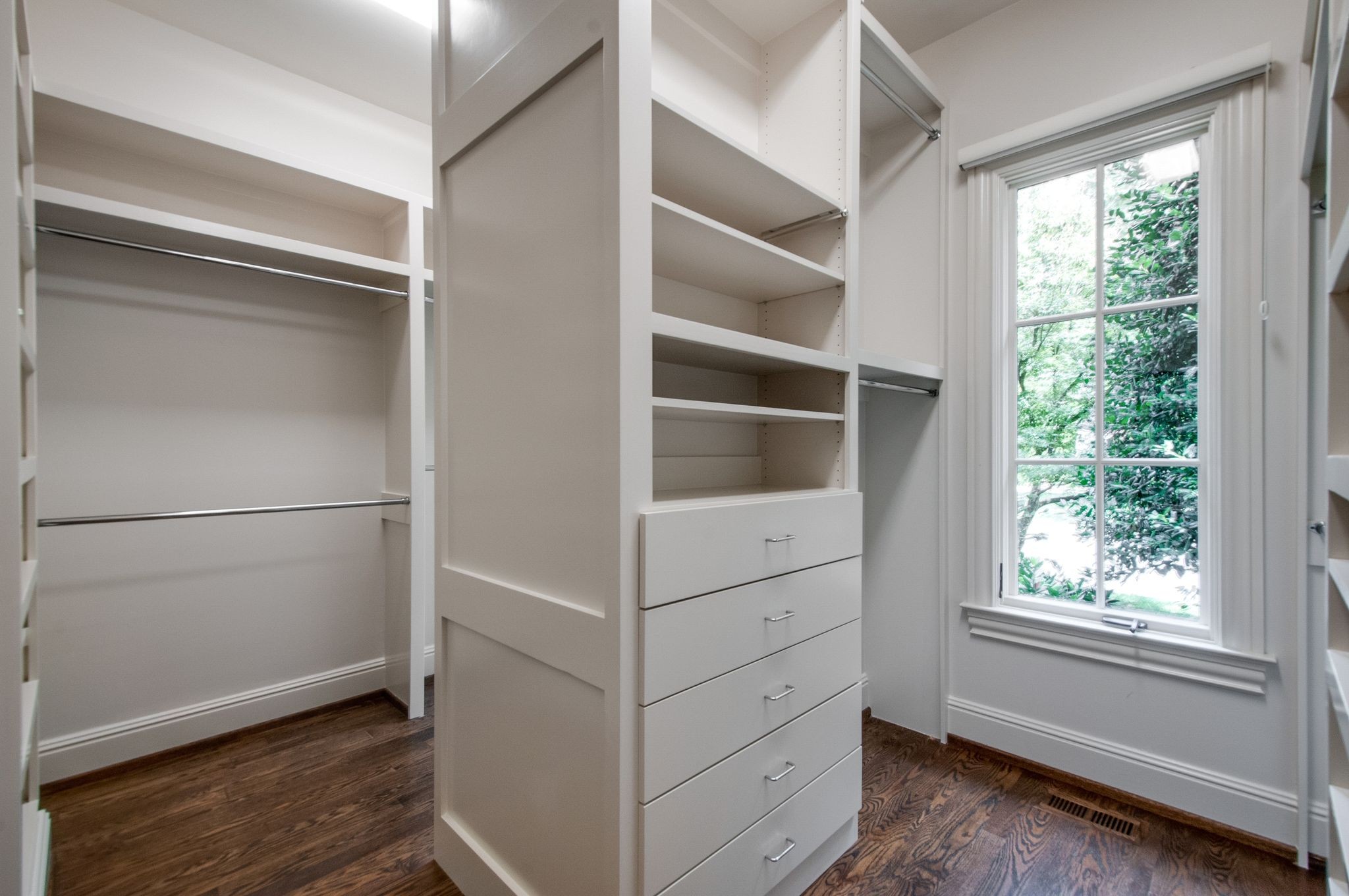 Walk-in closet with built-in white shelves and drawers beside a large window overlooking greenery.