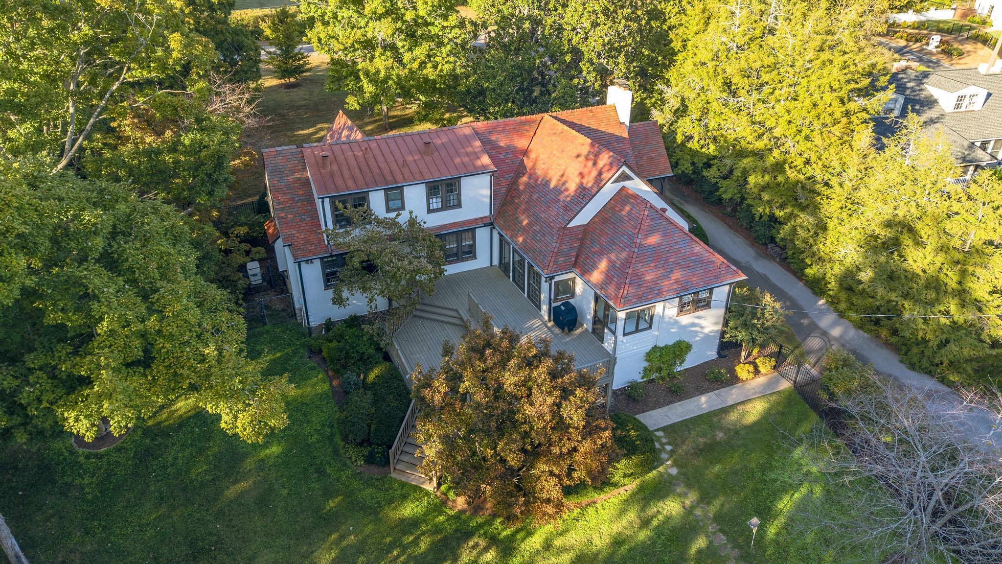 Aerial view of a white two-story home with red tile roofs, a broad deck, and a tree-lined lawn.