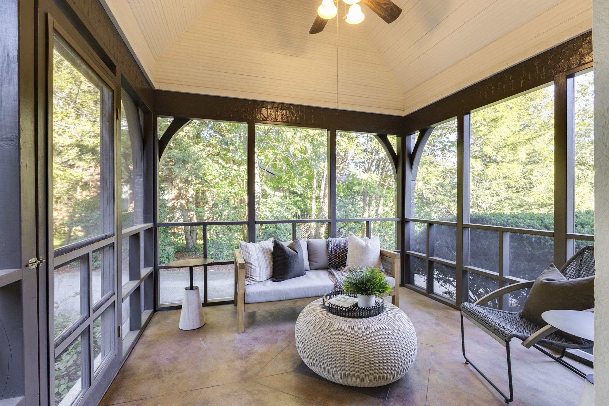 Furnished screened porch with vaulted ceiling, woven seating, and large windows overlooking trees.