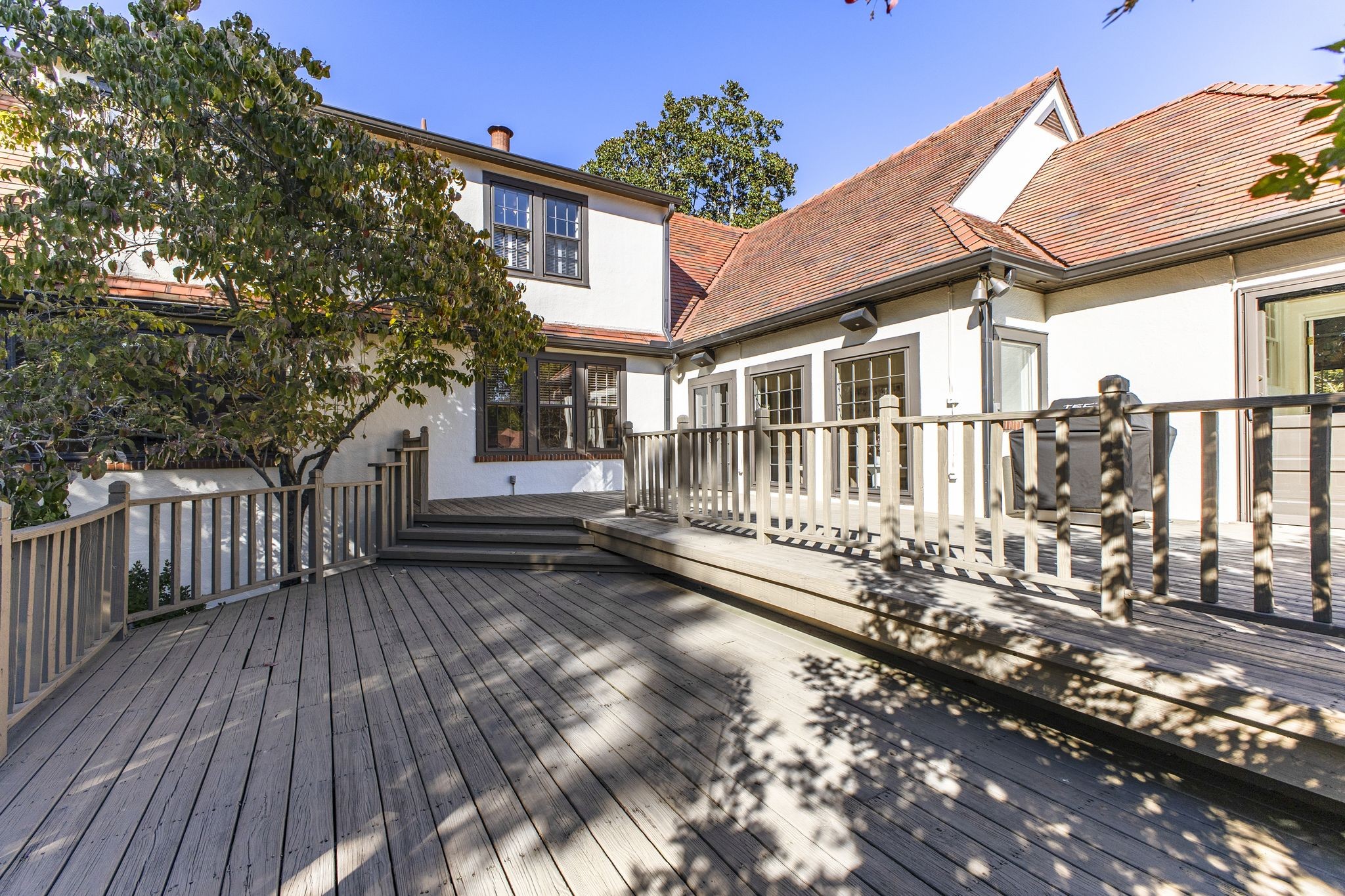 Rear deck with wood railings and steps beside a white house, shaded by a leafy tree.