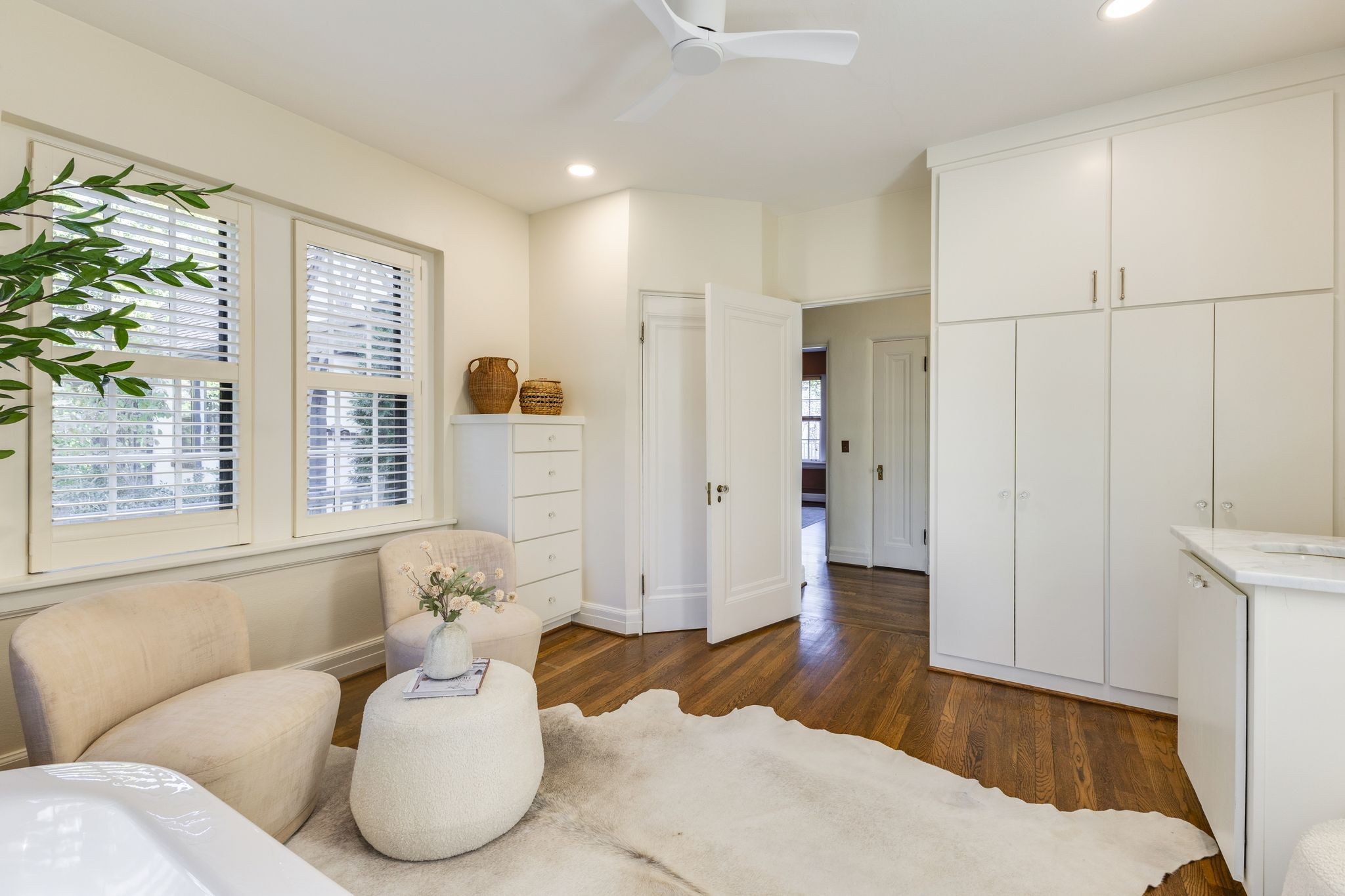 Bright bathroom with white built-in cabinets, shuttered windows, a small seating area, and dark hardwood floors.