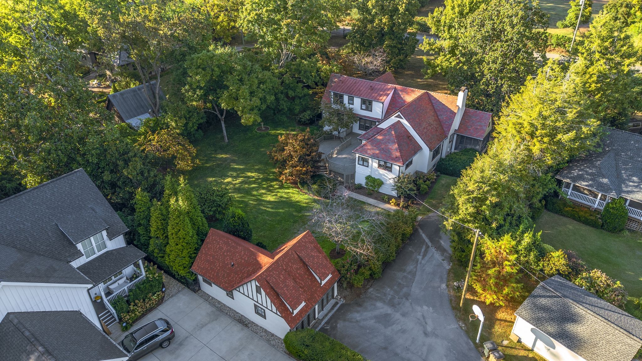 Aerial view of a white, red-roofed home amid mature trees, a broad lawn, and neighboring houses.