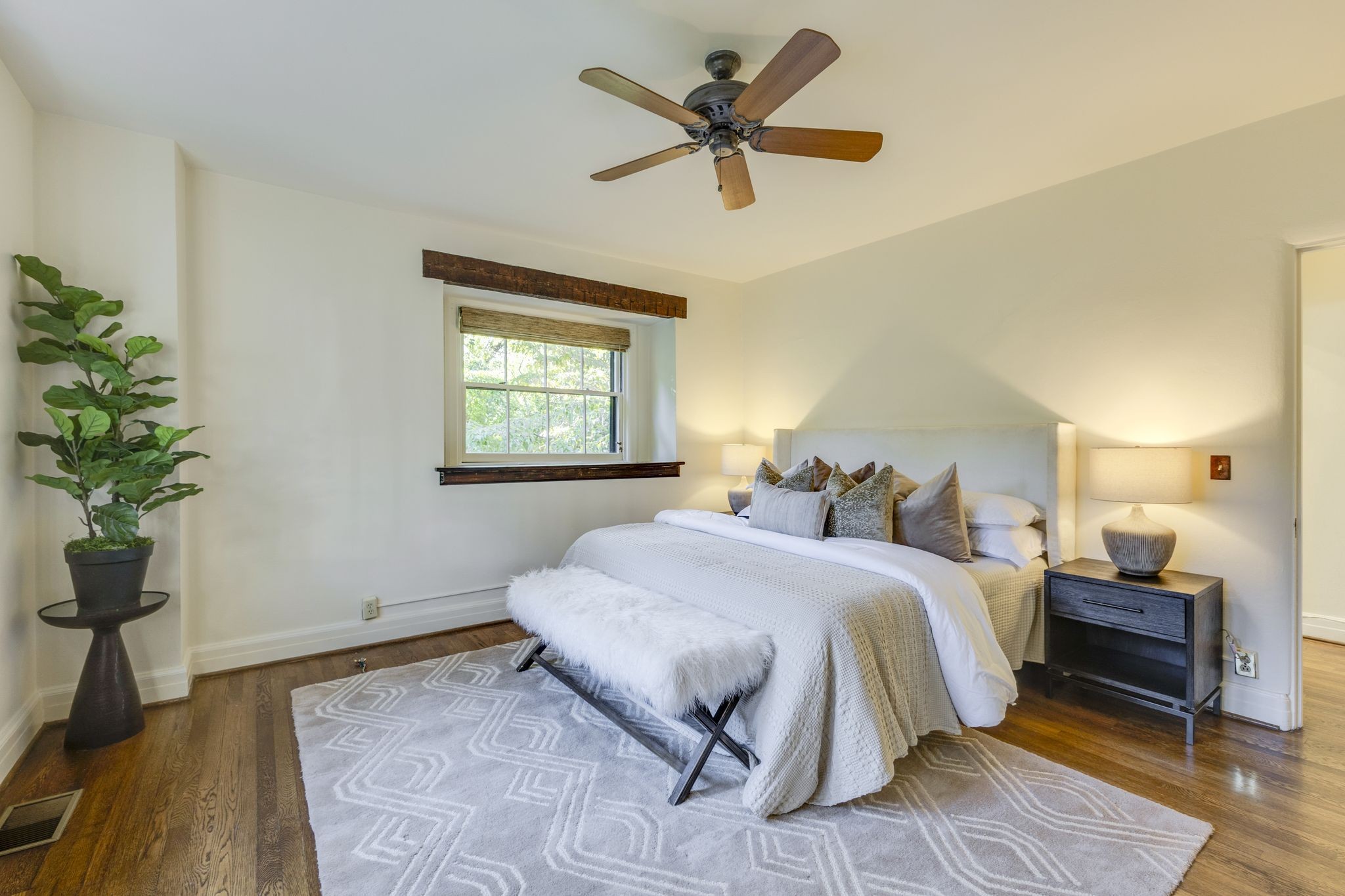 Staged bedroom with a white bed, faux-fur bench, hardwood floors, and a ceiling fan.