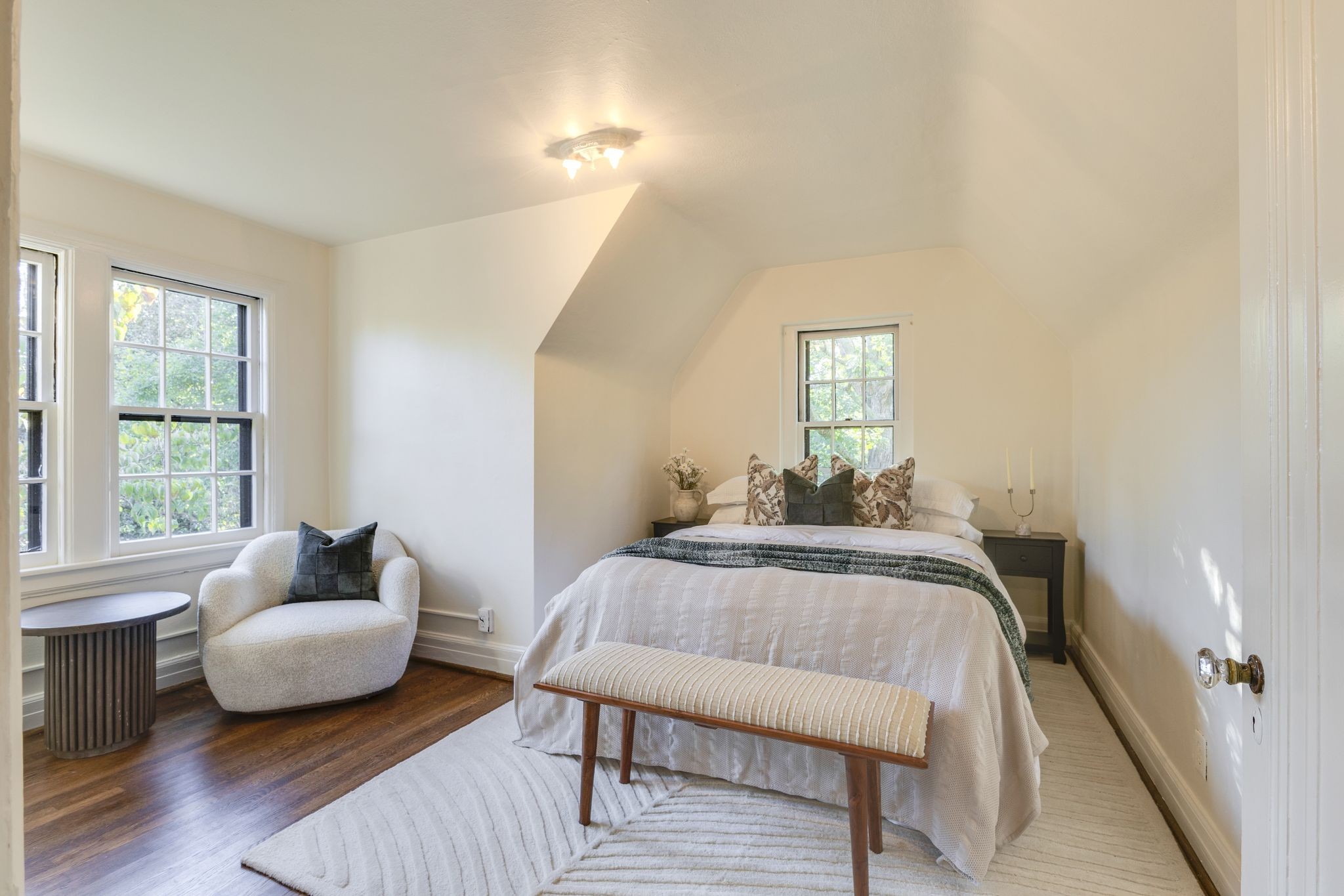 Bedroom with a white bed, sloped ceiling, window-side armchair, and dark wood floors.