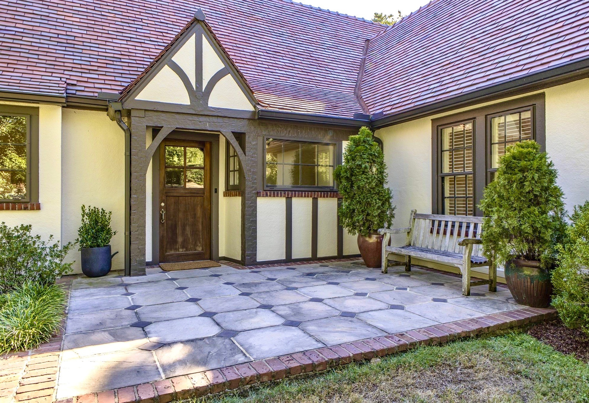 Home entrance with a timber-trimmed doorway, stone-paved courtyard, potted evergreens, and wooden bench.