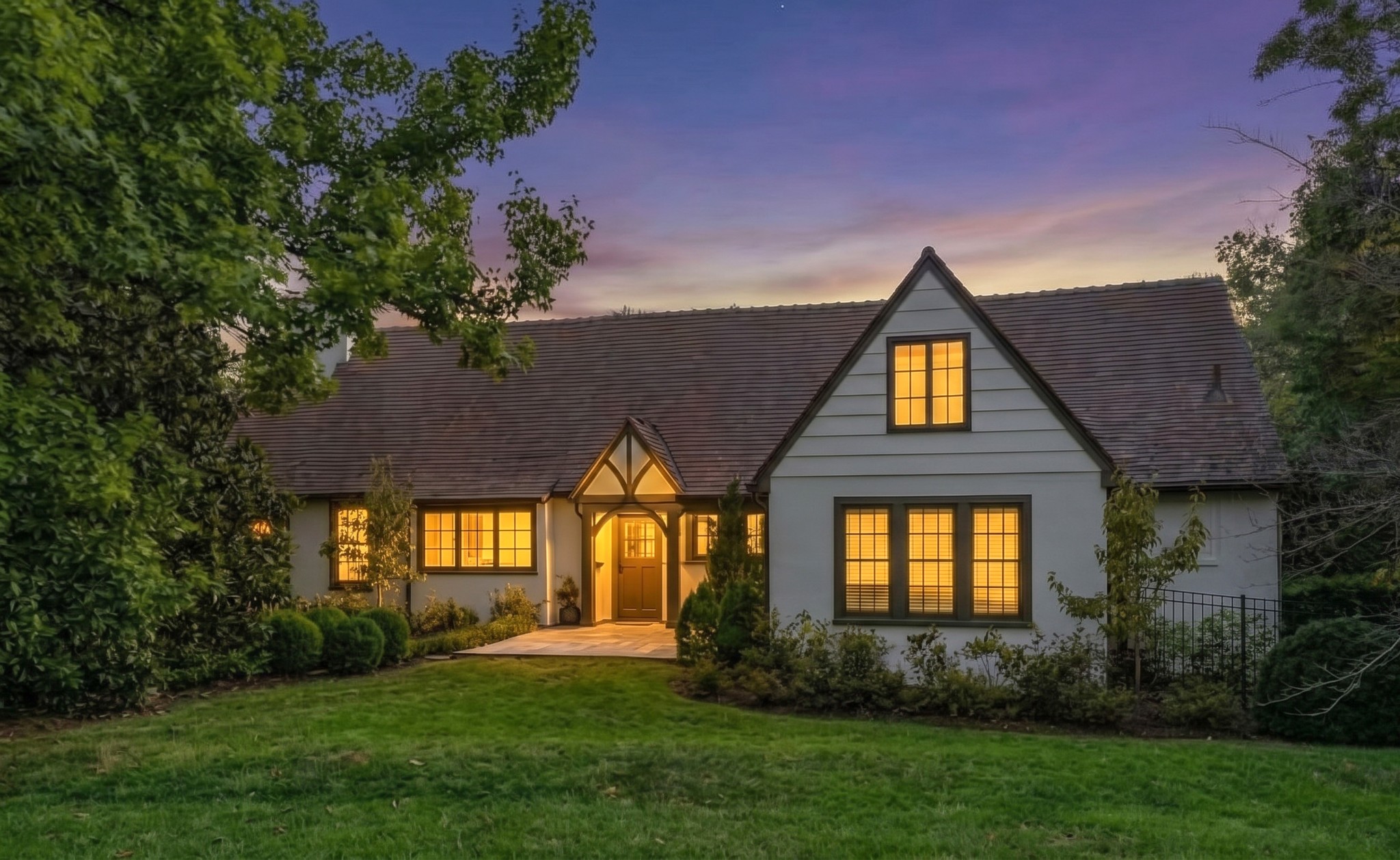 House at dusk with a timber-trimmed entry, glowing windows, and landscaped lawn.