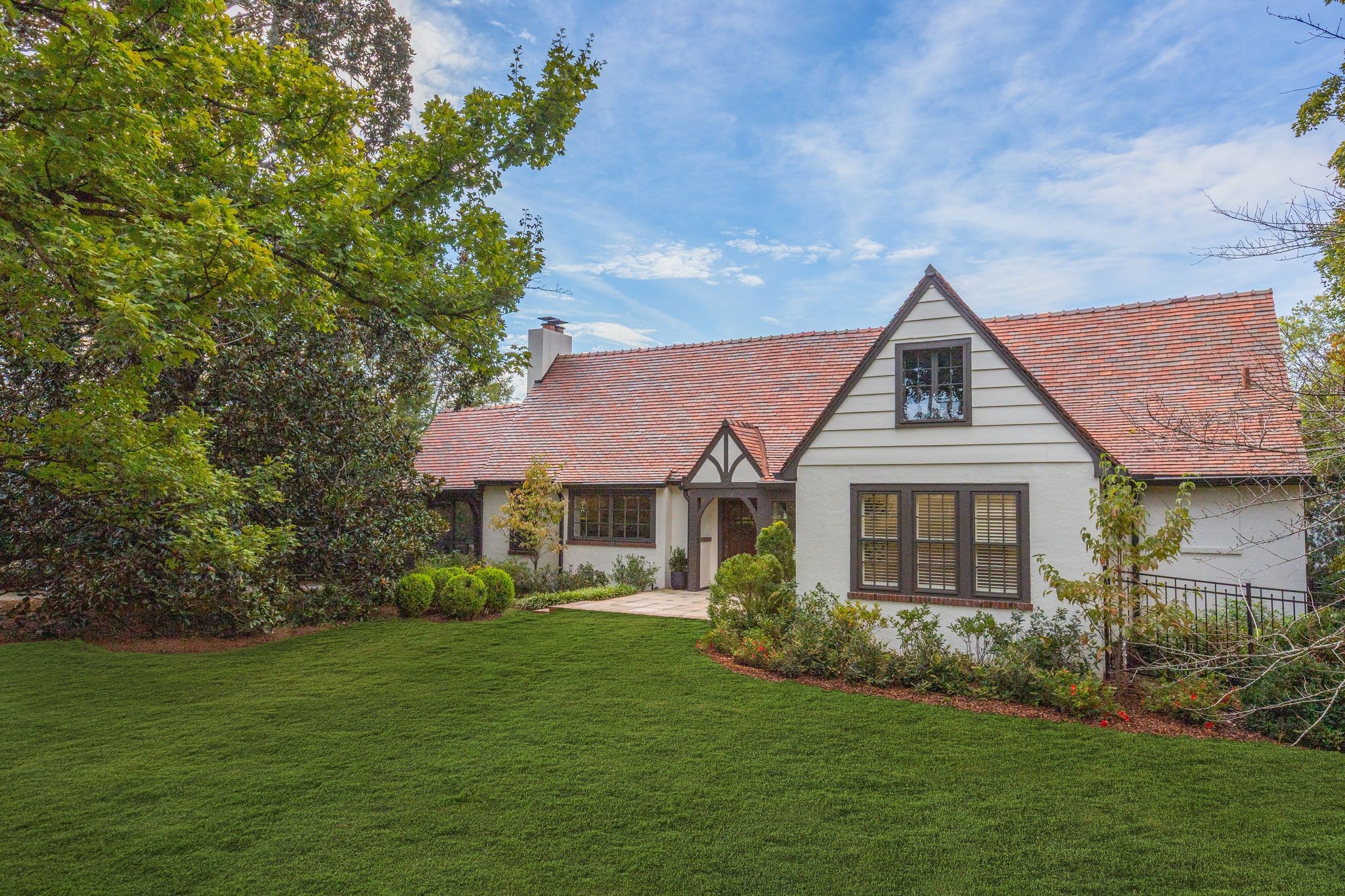 White Tudor-style home with a steep red tile roof, landscaped shrubs, and a broad green lawn.