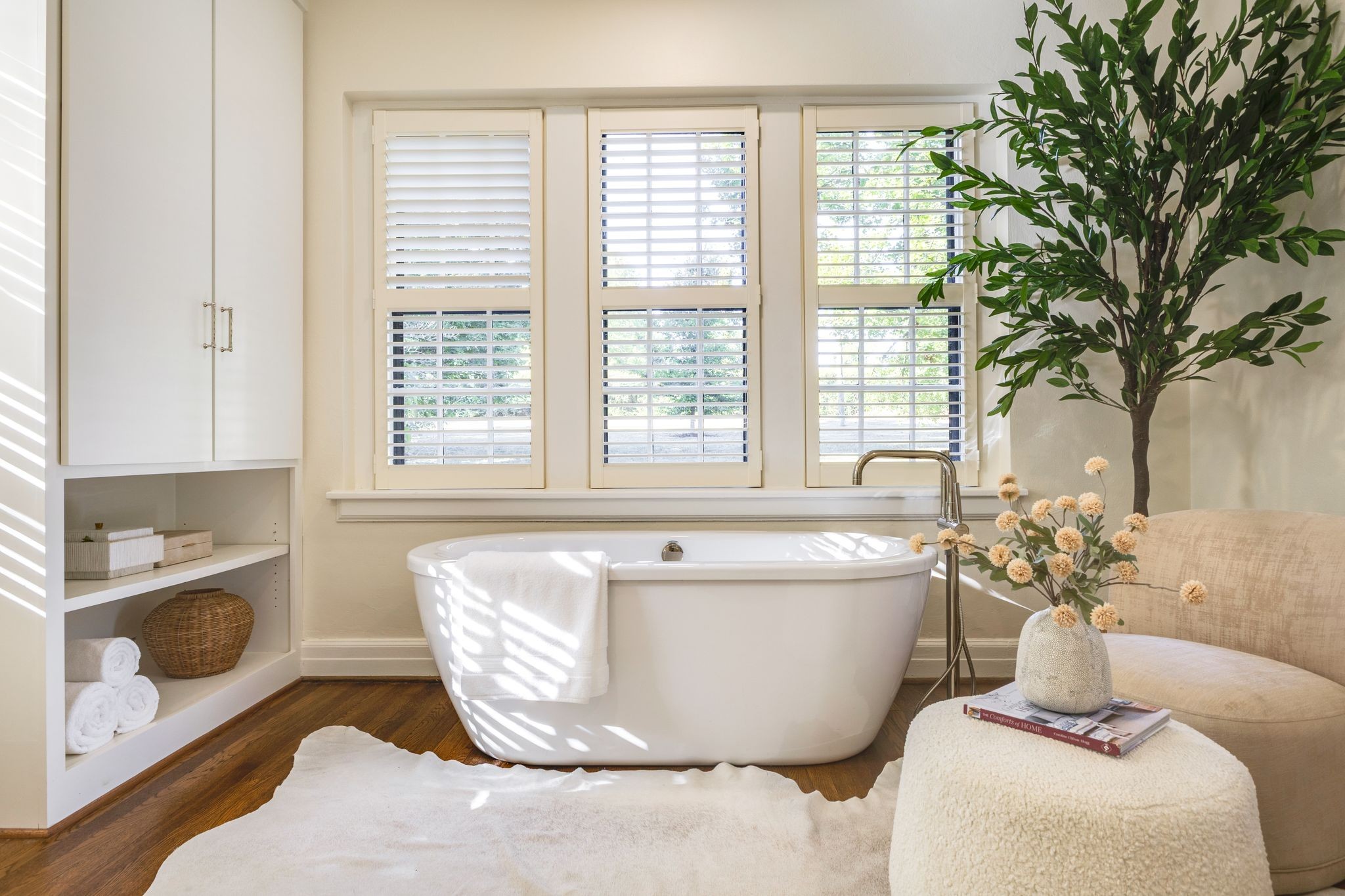 Bright bathroom with a freestanding soaking tub beneath shuttered windows and built-in towel shelves.