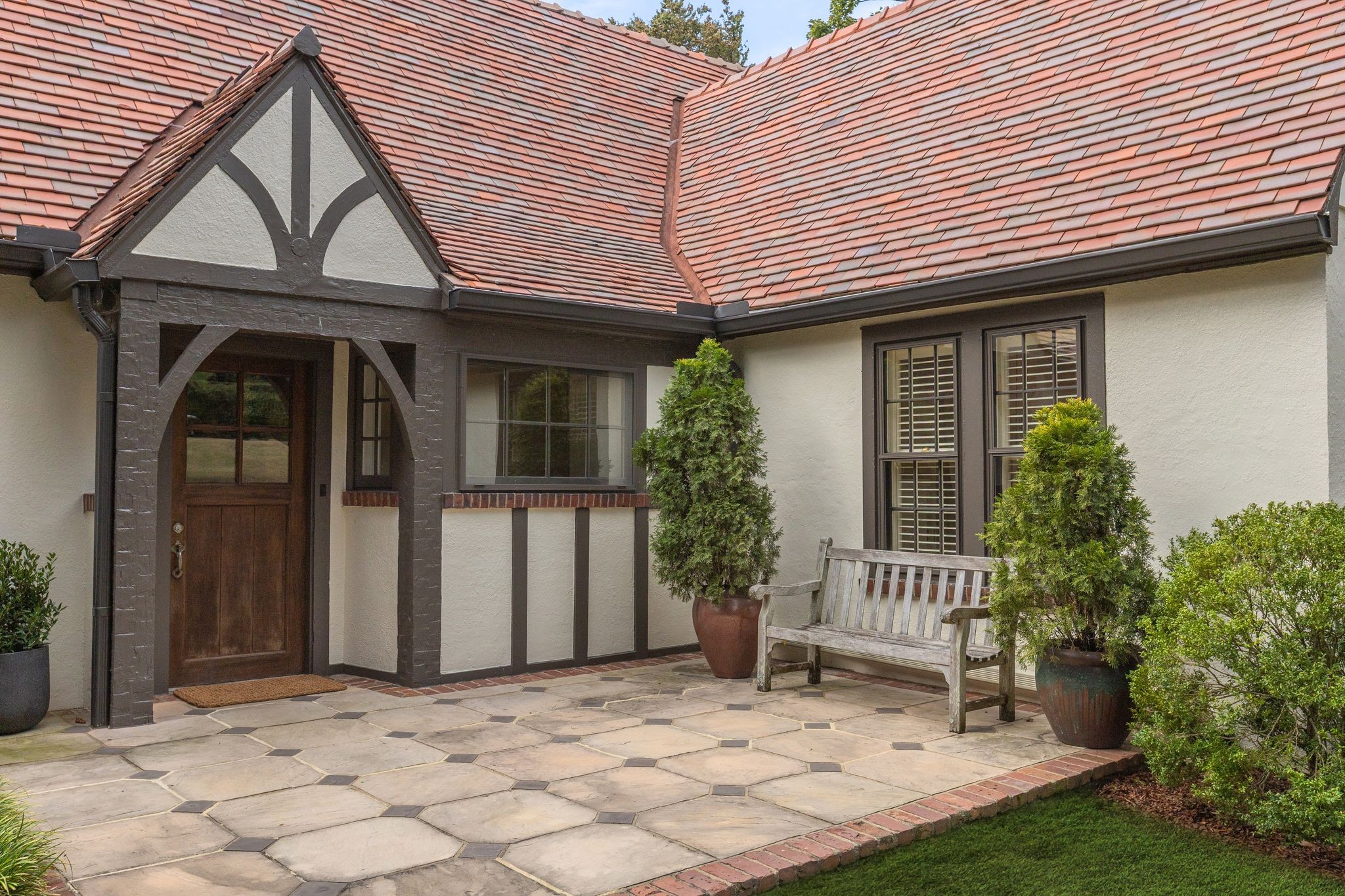 Home entrance with red tile roof, timber-trimmed gable, stone courtyard, potted shrubs, and wooden bench.