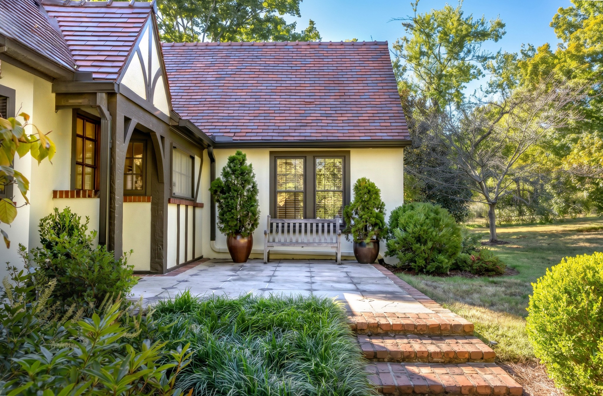 Cream home with a red tiled roof, stone patio, brick steps, and a bench between potted evergreens.