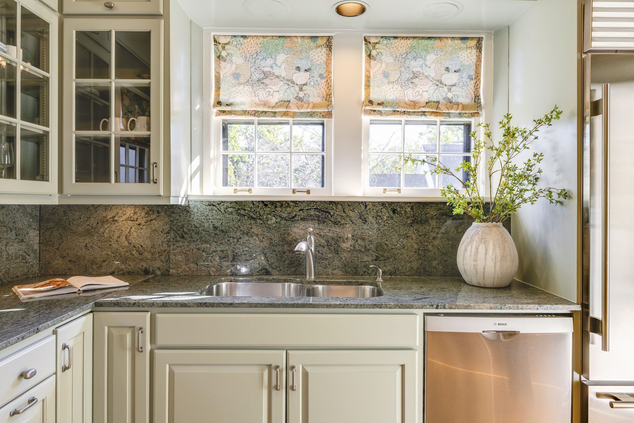Kitchen sink beneath two floral Roman shades, framed by dark granite counters and cream cabinetry.