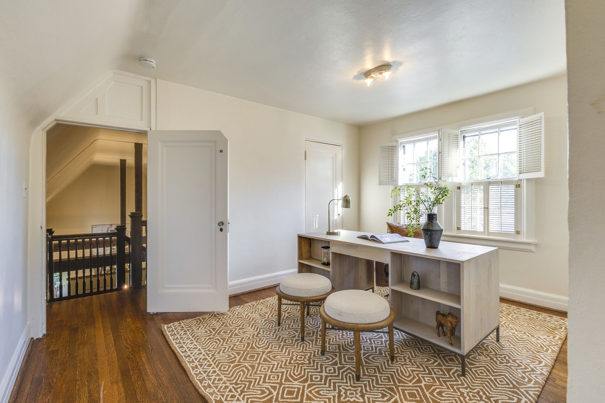 Staged home office with a light wood desk, patterned rug, and shuttered windows beside an open stairway.