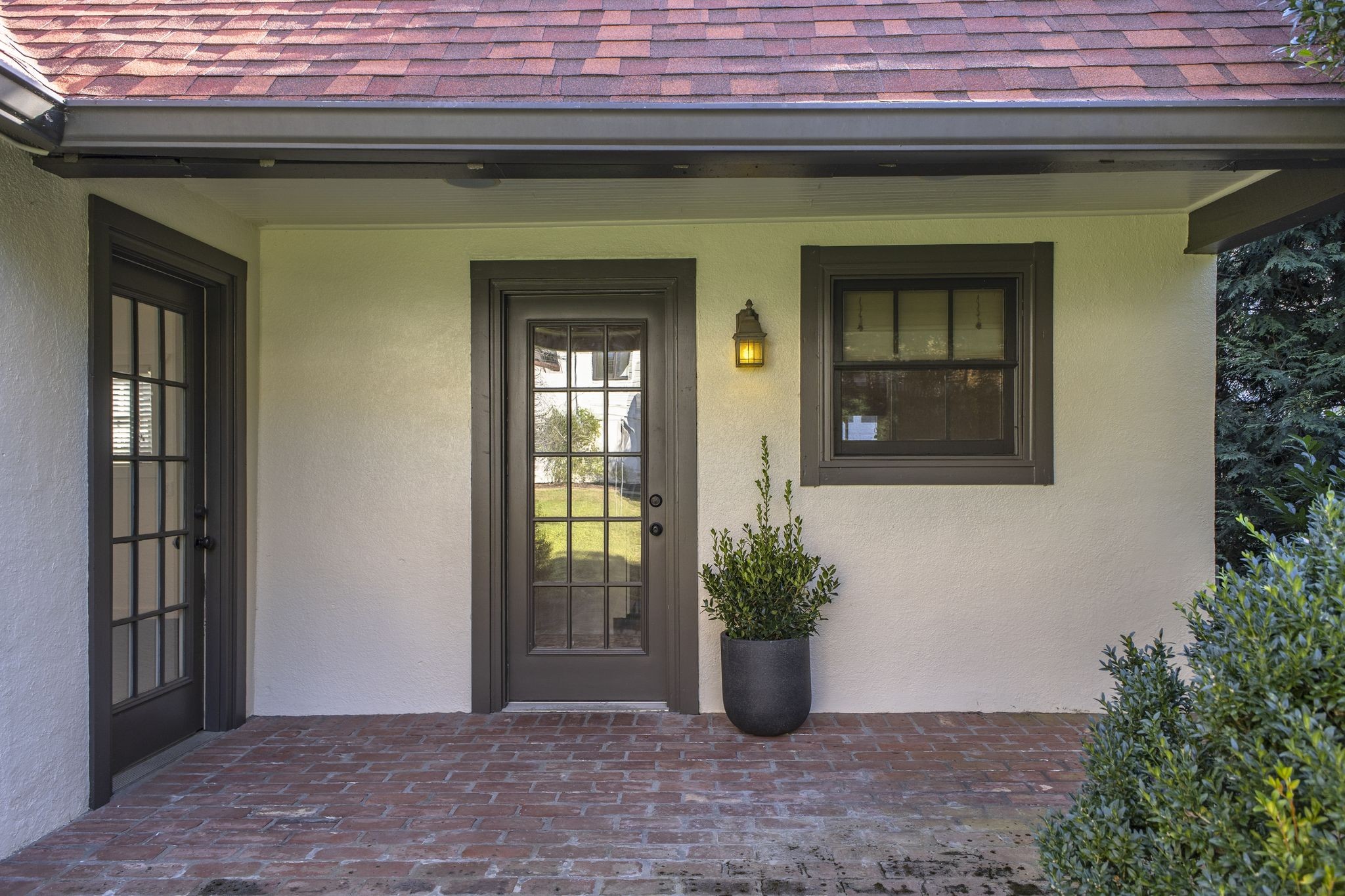Covered front entry with dark-framed glass doors, brick flooring, and a potted shrub.