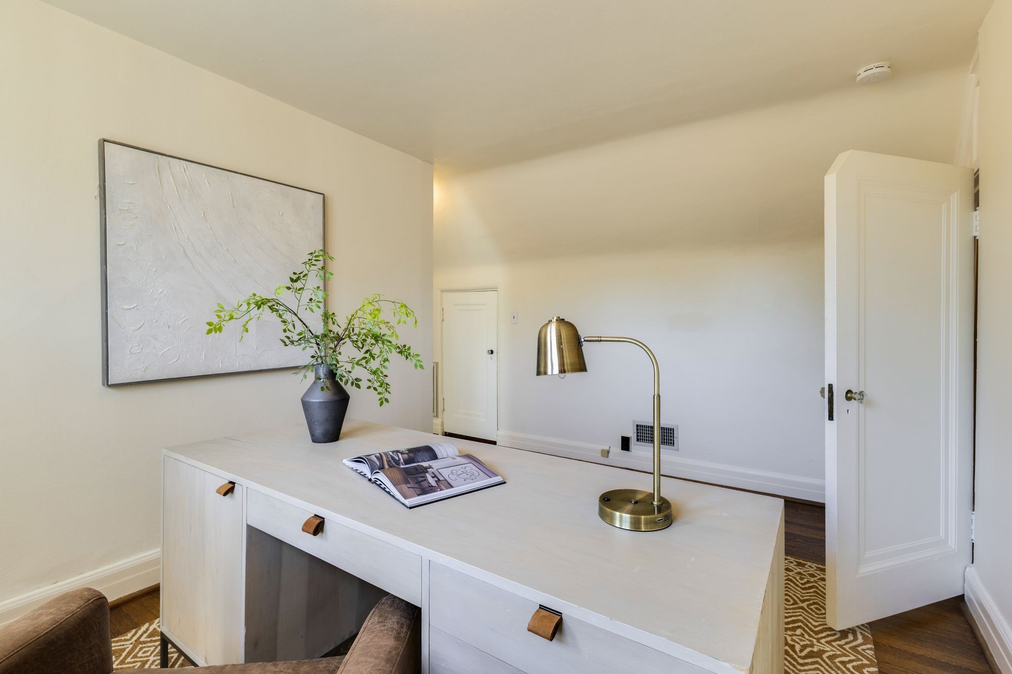 Furnished home office with a white desk, brass lamp, leafy plant, and large abstract wall art.