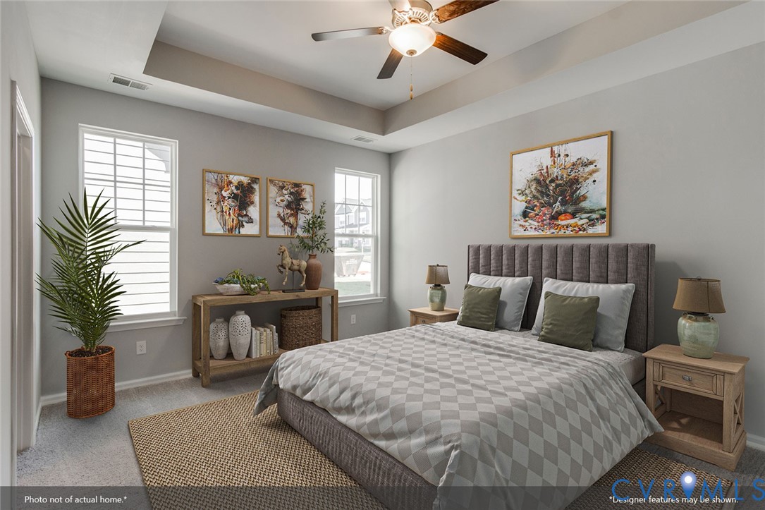 Staged bedroom with a large bed, gray upholstered headboard, tray ceiling, and ceiling fan.