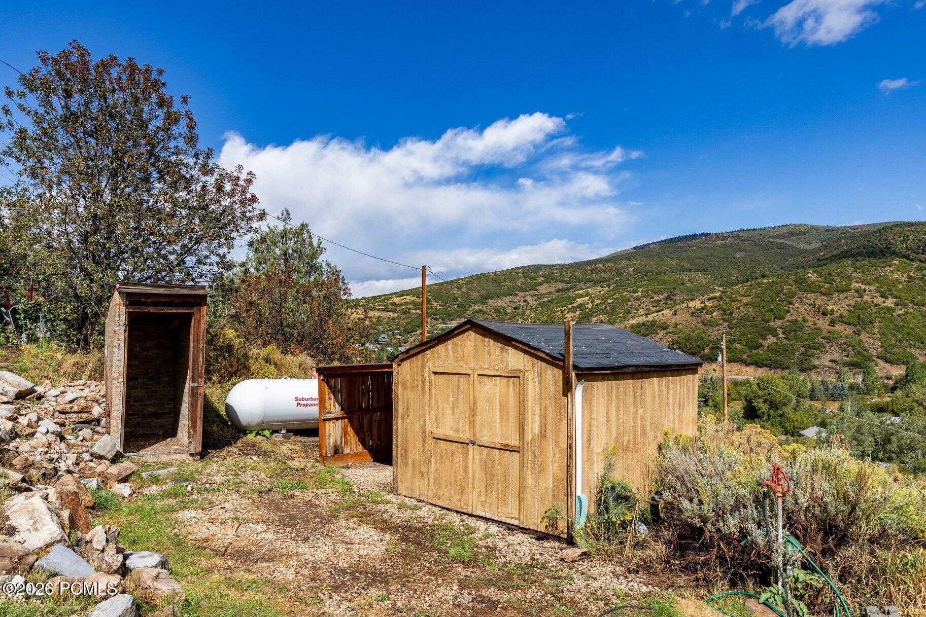 Wooden storage sheds and a propane tank sit on a rocky hillside below scrub-covered mountains.