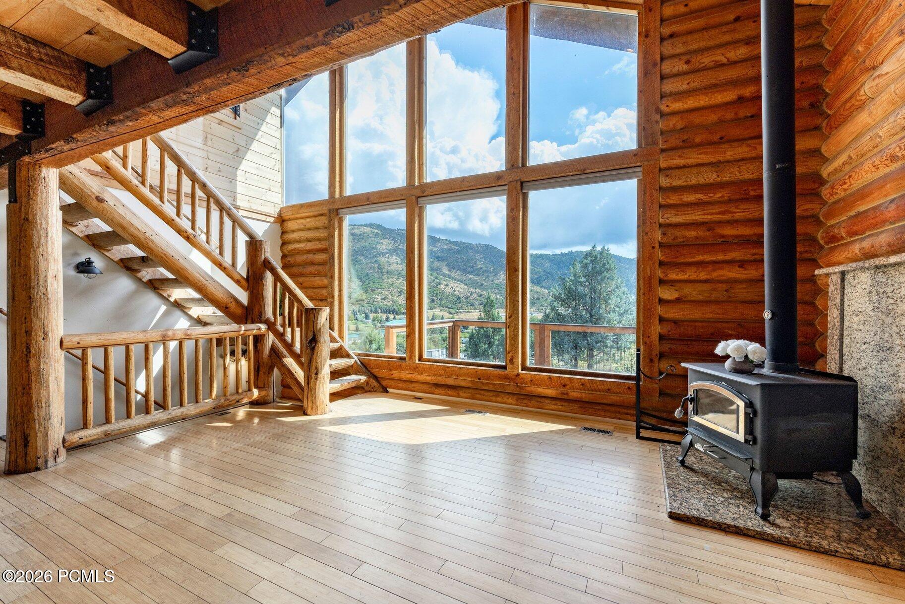 Log-cabin living room with a tall mountain-view window, exposed timber staircase, and wood-burning stove.
