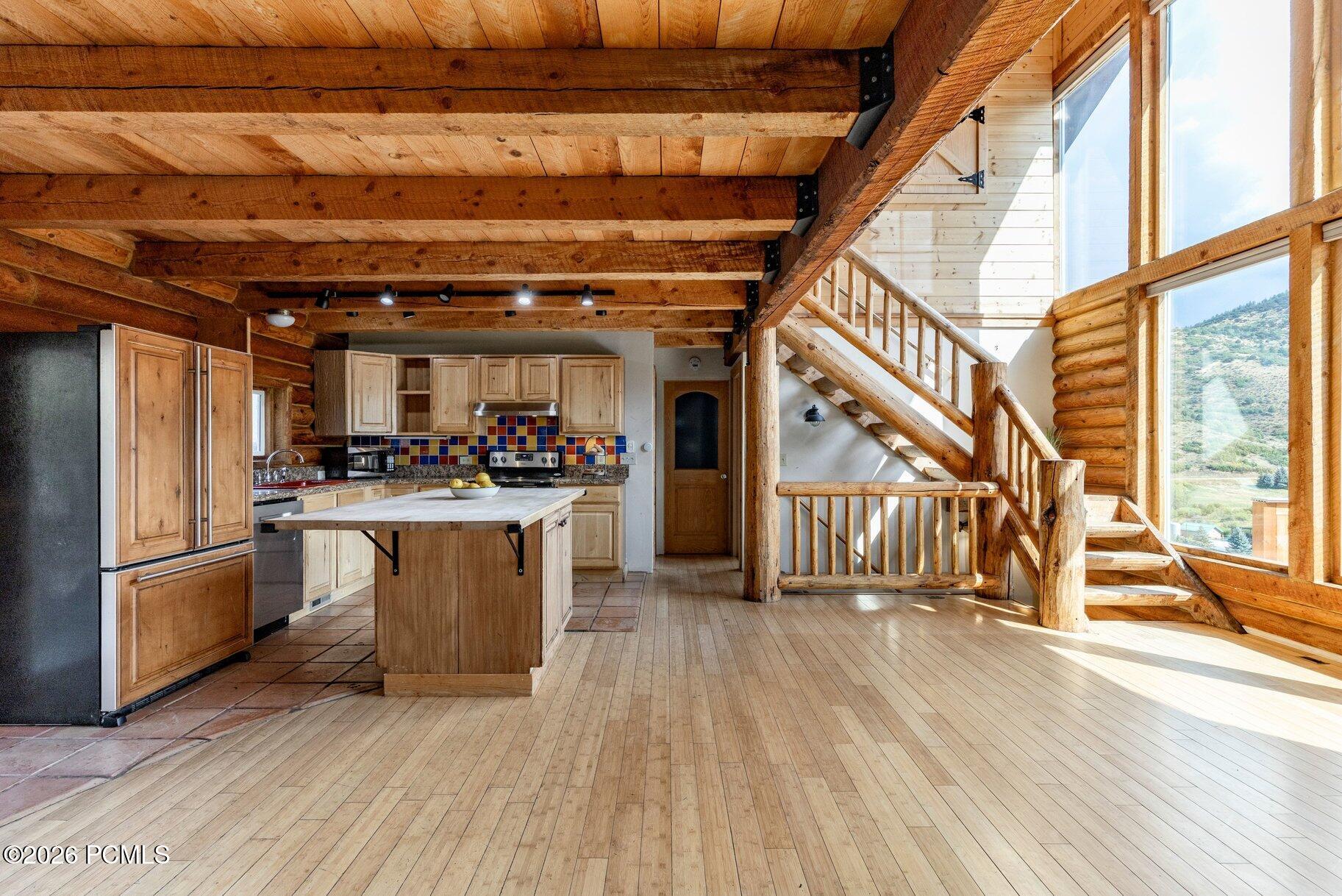 Rustic kitchen and open living area with exposed timber beams, log staircase, and mountain views through tall windows.