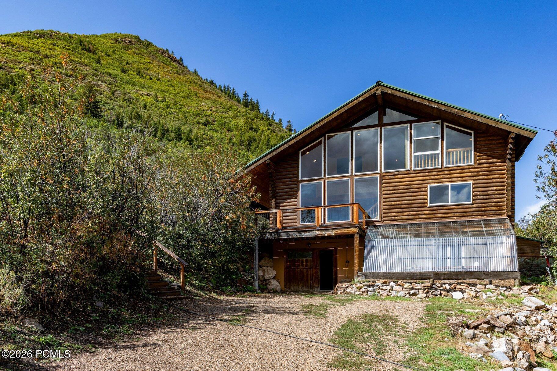 Log home with large windows and an adjoining greenhouse beneath a wooded hillside.