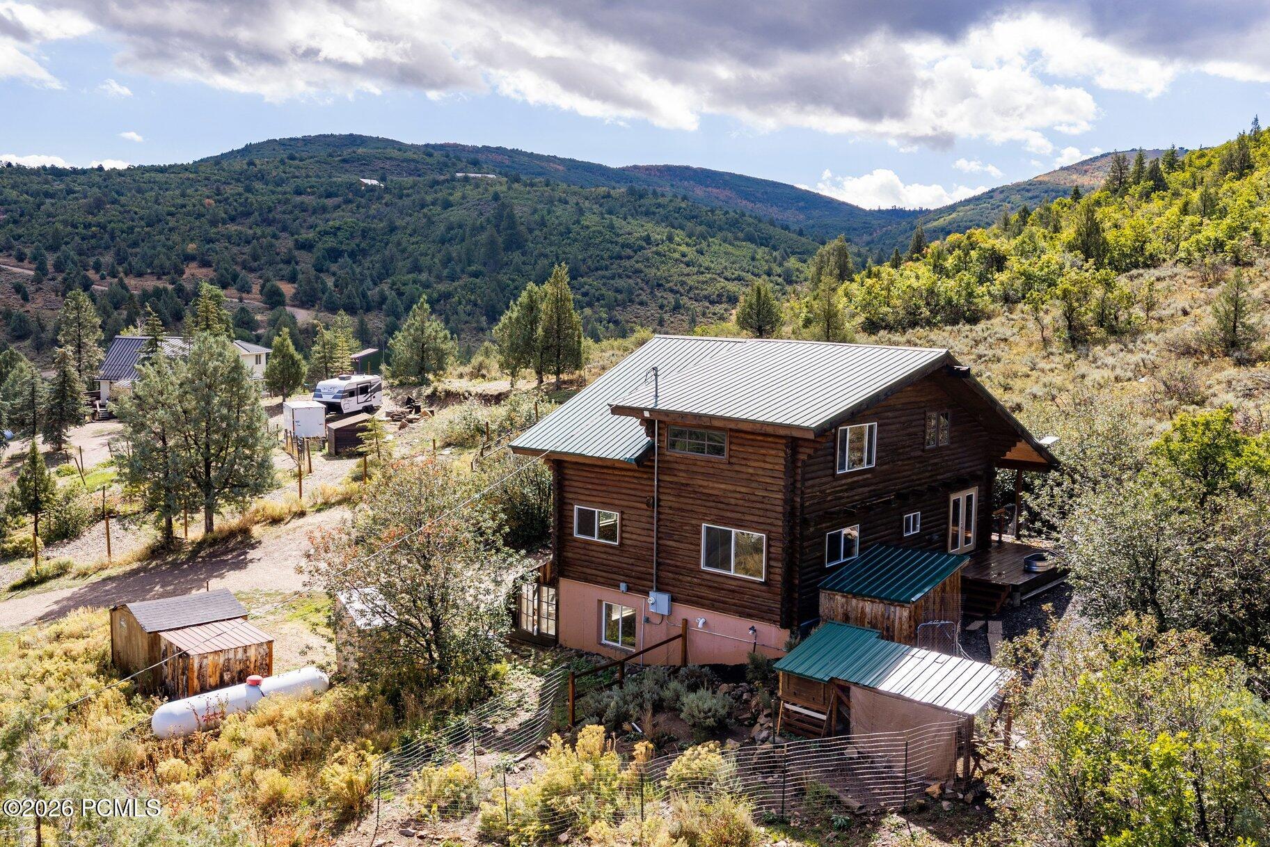 Elevated view of a log-sided home with a metal roof among wooded hills and outbuildings.