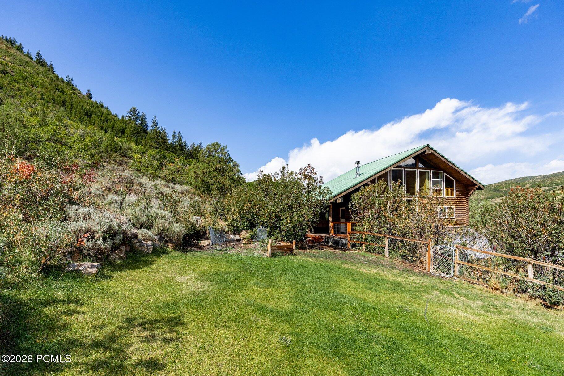 Log cabin with a green roof in a grassy yard, surrounded by wooded hills and a fenced garden.