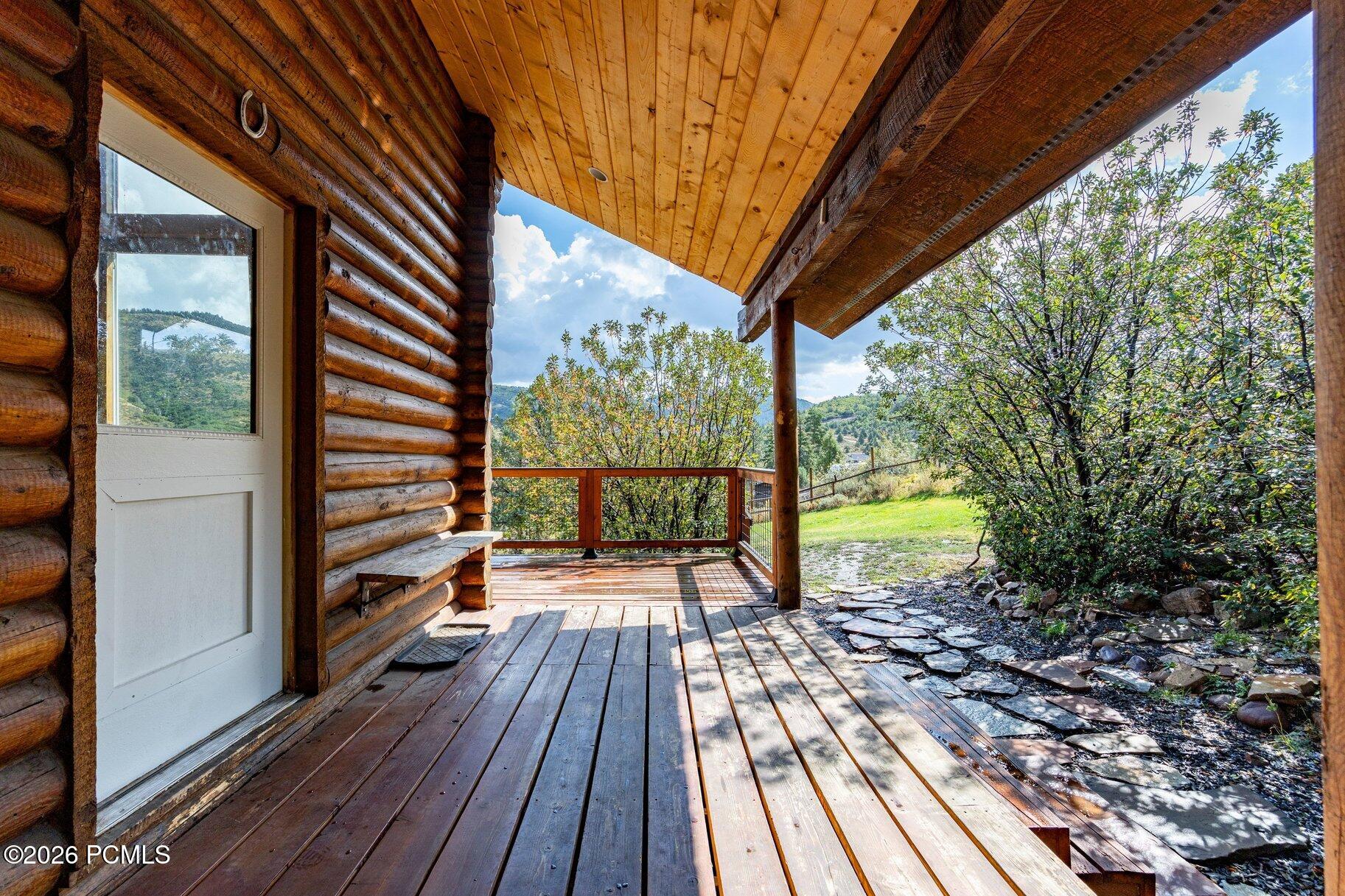 Covered log-home deck overlooking leafy trees, a grassy yard, and green hills.