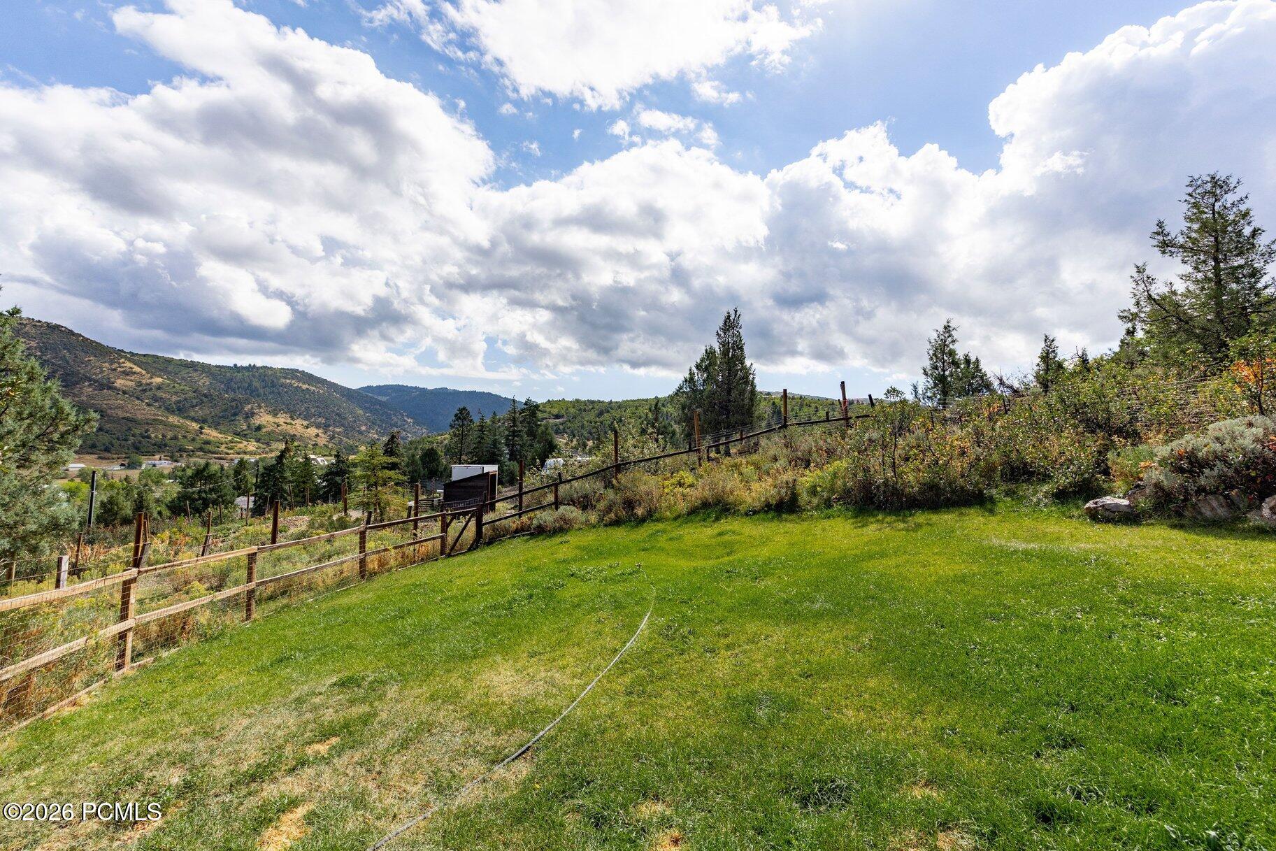 Sloping green lawn and fenced hillside overlooking a wooded valley and distant mountains.