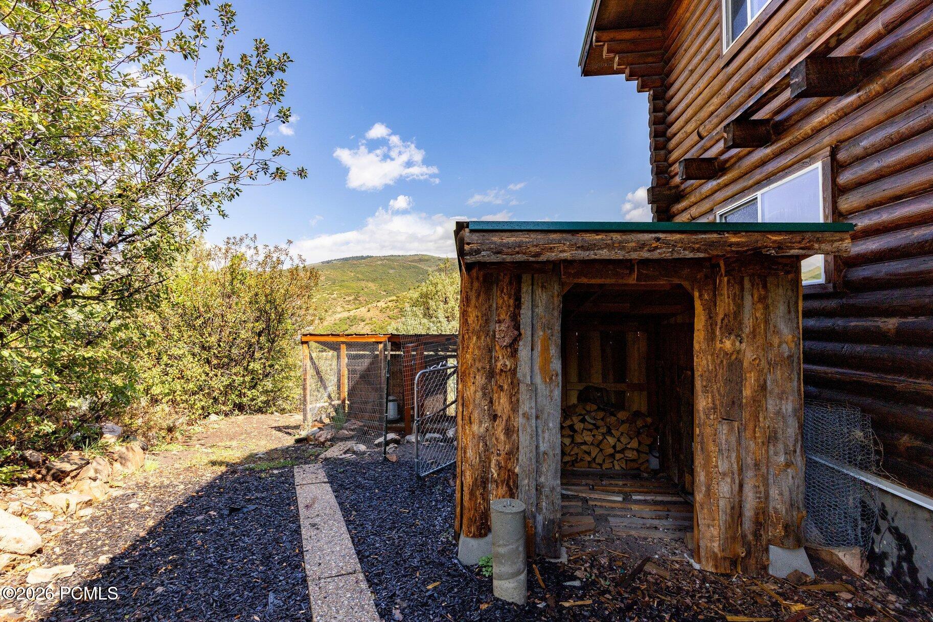 Log home exterior with covered firewood storage, a fenced enclosure, and a green hillside beyond.