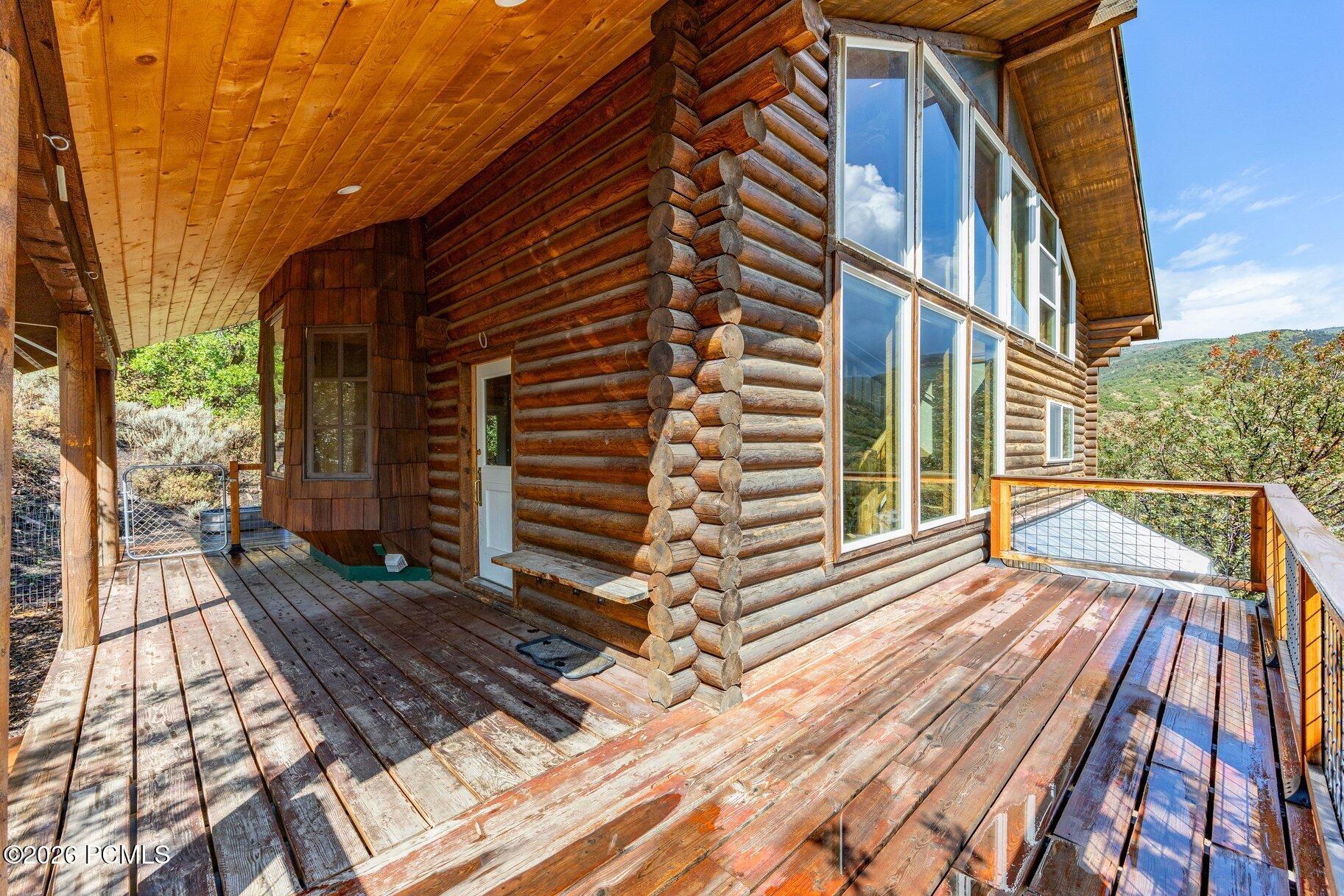 Log home with a broad weathered wood deck, covered porch, and tall windows overlooking a wooded hillside.