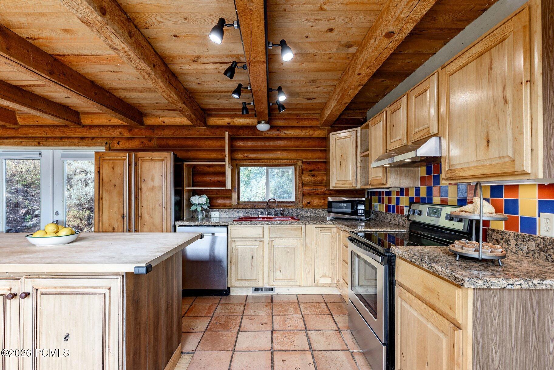Kitchen with pale wood cabinetry, colorful tile backsplash, granite counters, and a rustic log ceiling.
