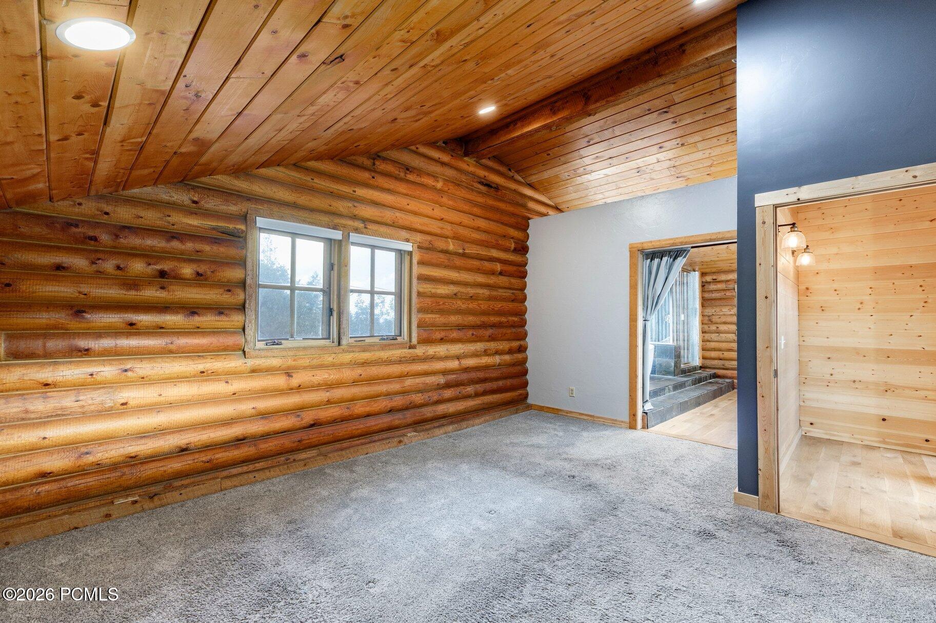 Empty carpeted room with log walls, a sloped wood ceiling, and a doorway to a wood-paneled room.