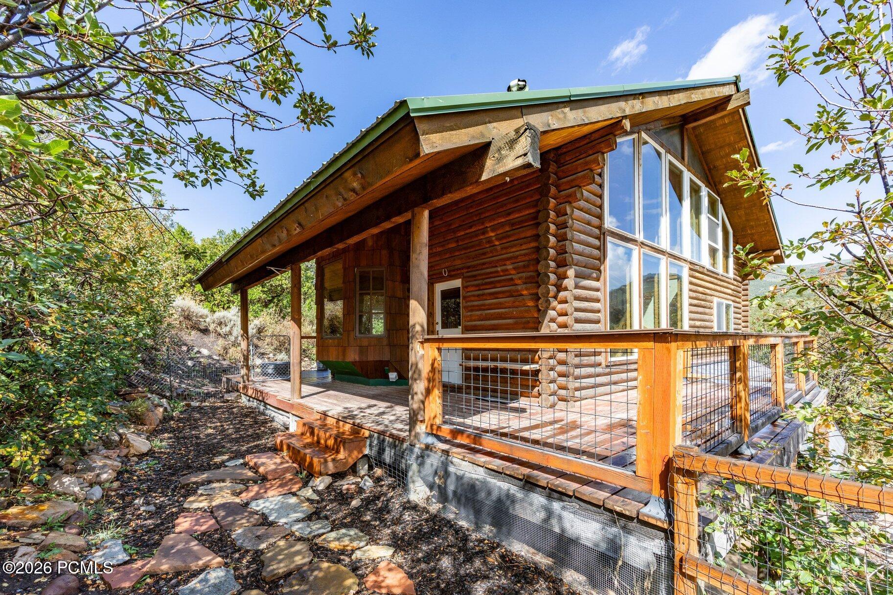 Log cabin with a raised deck, large windows, and stone steps among leafy trees.